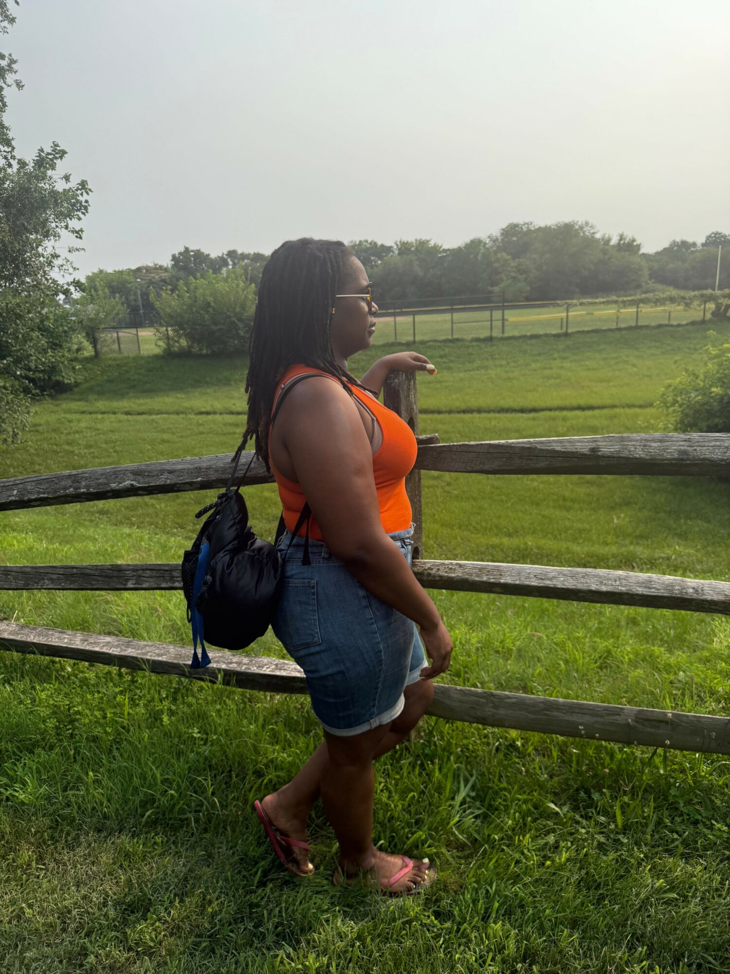 Person with long hair, wearing an orange top and denim shorts, standing on grass near a wooden fence.