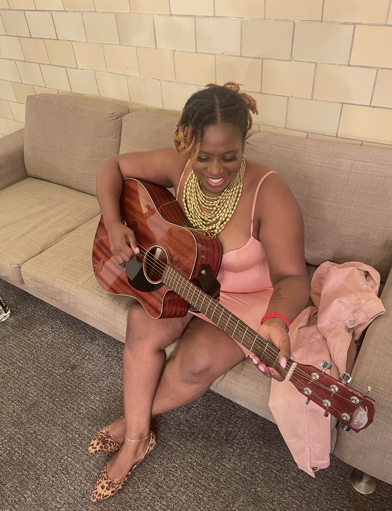 Woman sitting on a sofa playing an acoustic guitar, smiling, wearing a pink dress and leopard print shoes.