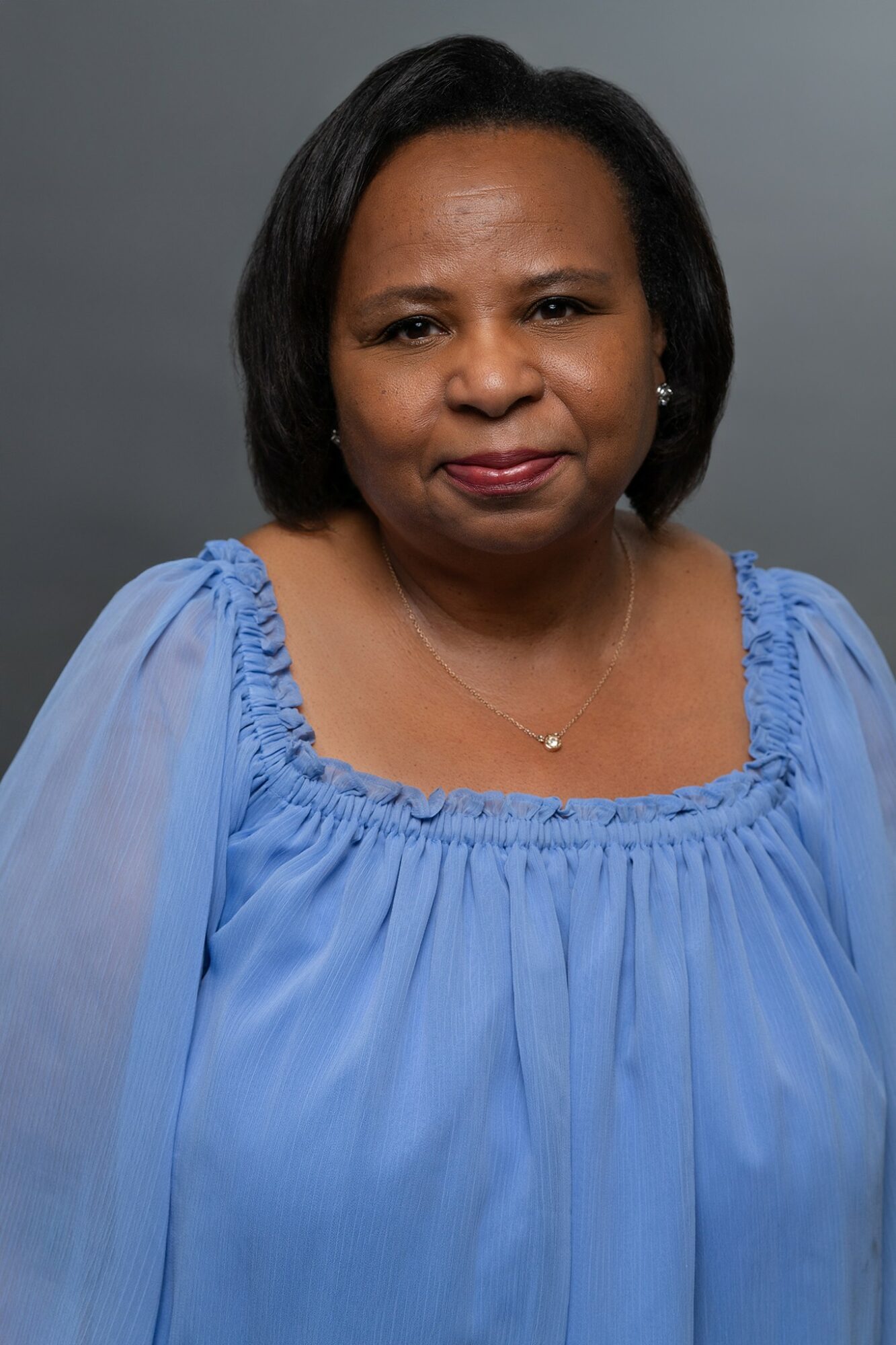 A woman with shoulder-length black hair wearing a blue blouse with ruffled neckline, looking at the camera against a gray background.