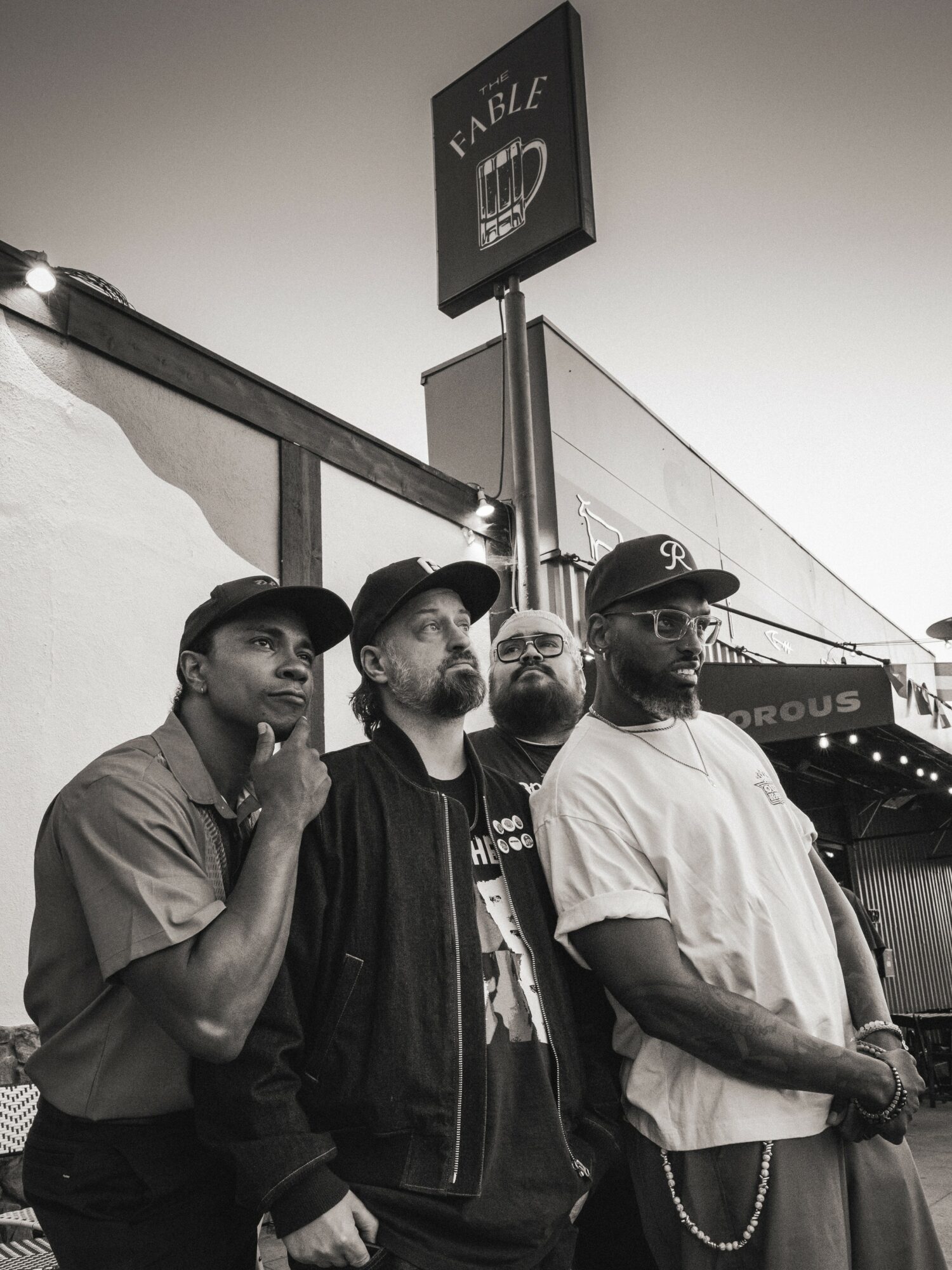 Four men standing outdoors near a building with a sign above them, in black and white.