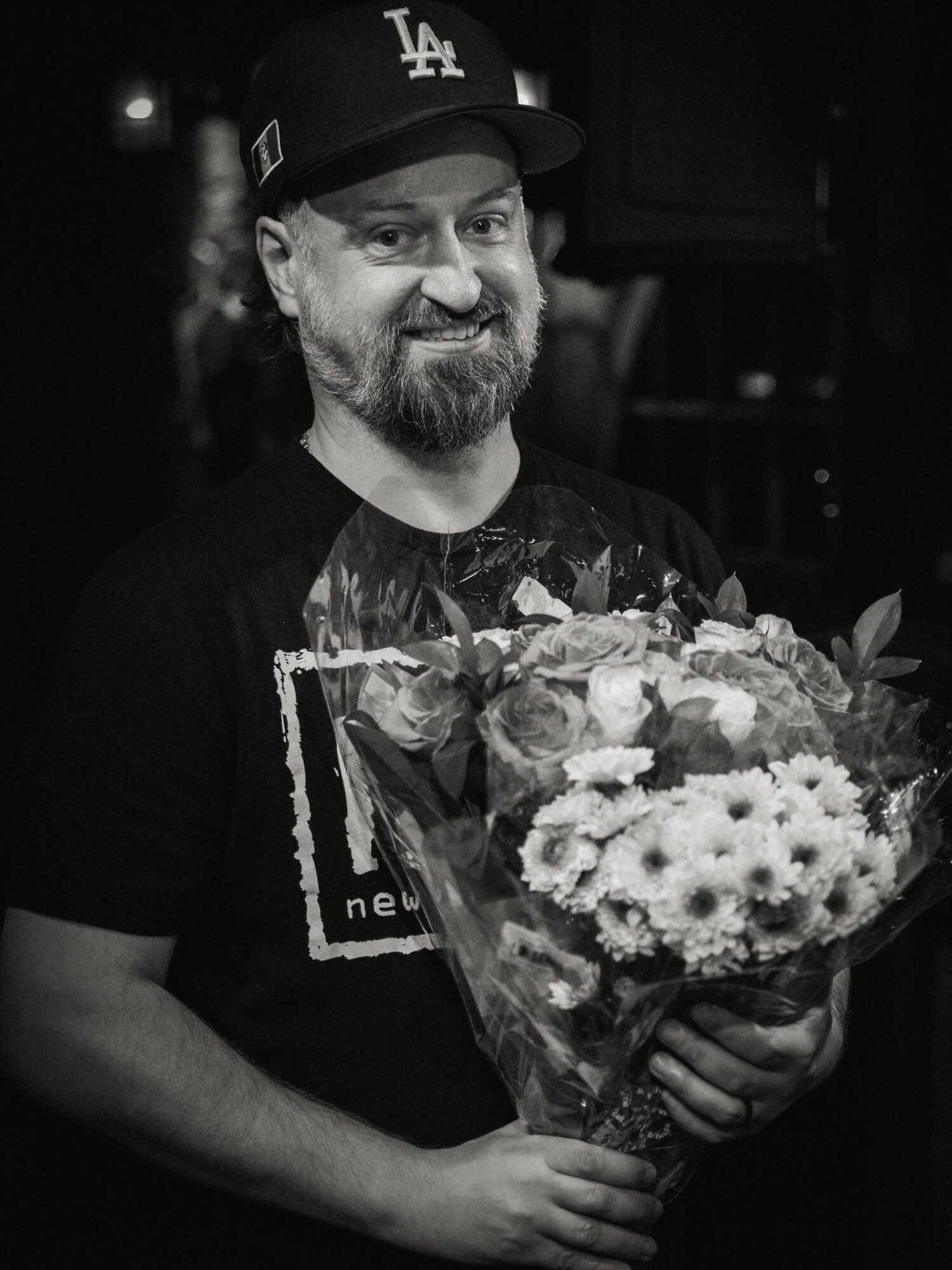 Man with a beard wearing a Los Angeles cap holding a bouquet of flowers, smiling.