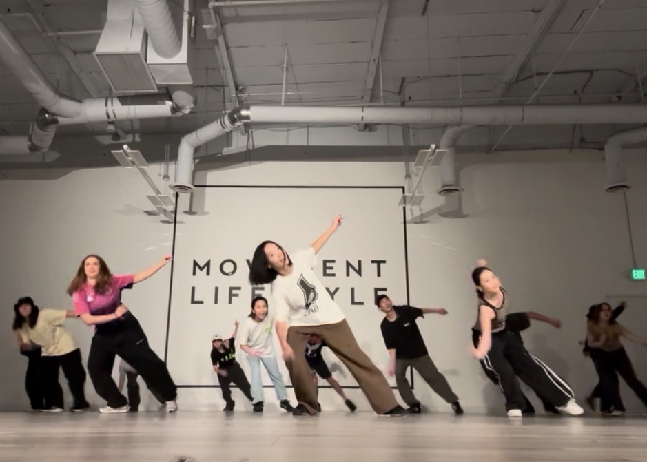 Group of people dancing in a studio with a white wall and industrial ceiling, some with arms raised or extended.