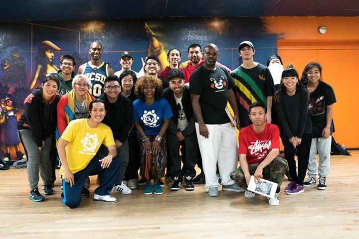 Group of diverse people posing indoors with a colorful mural background, some smiling and holding items.