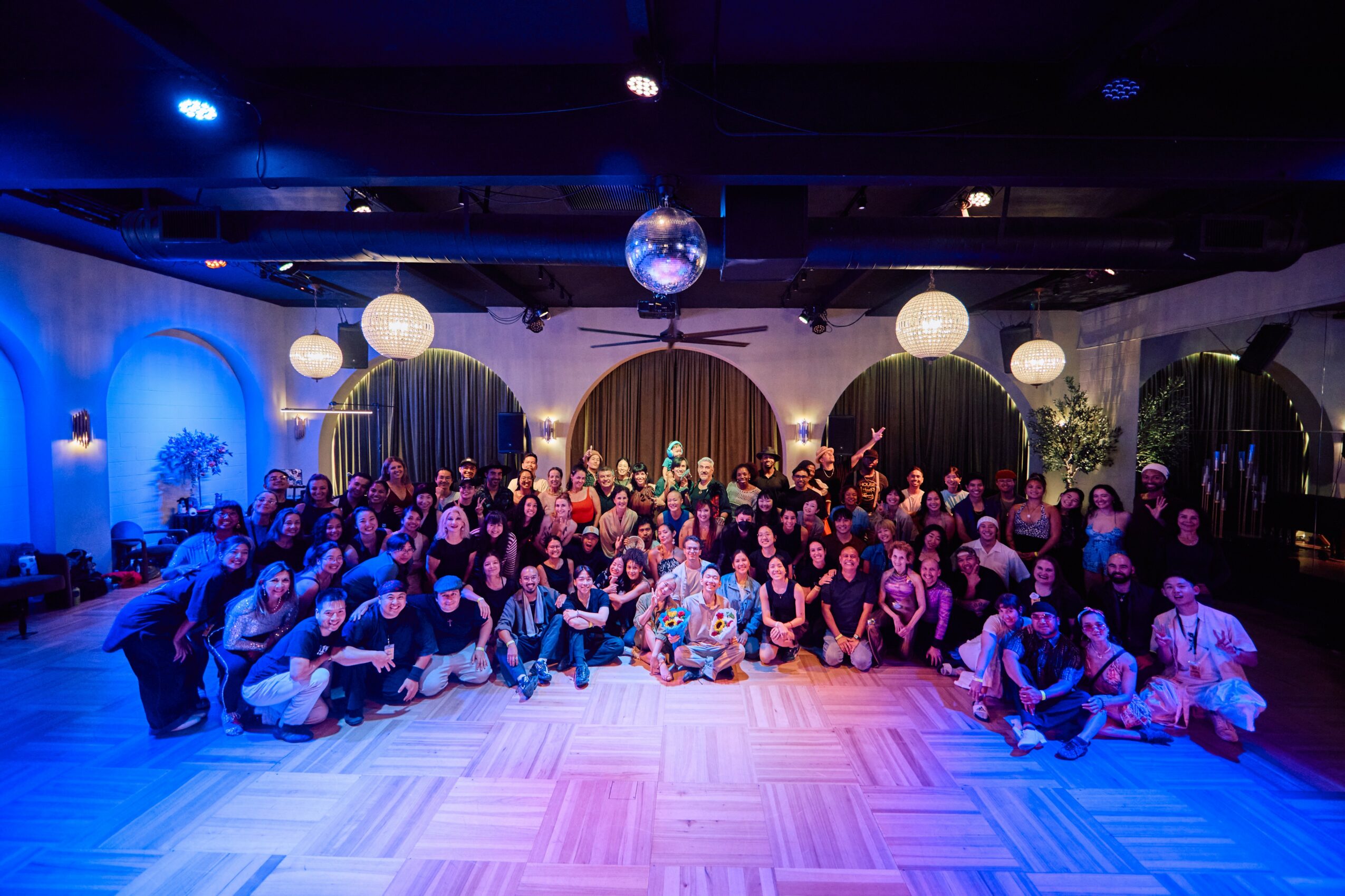 Large group of people gathered in a decorated indoor venue with curtains and hanging lights, posing for a group photo.