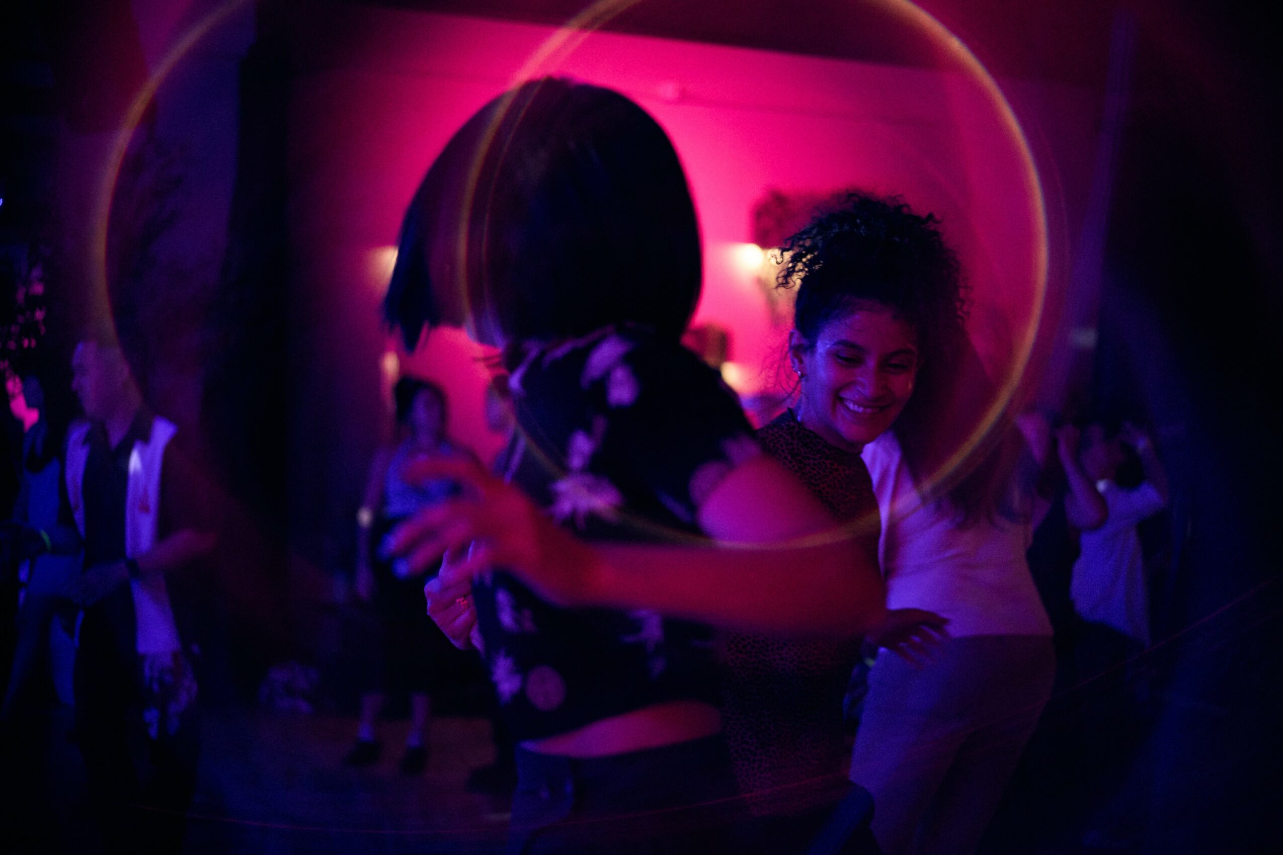 Two women hugging and smiling in a dimly lit, colorful party setting with other people in the background.