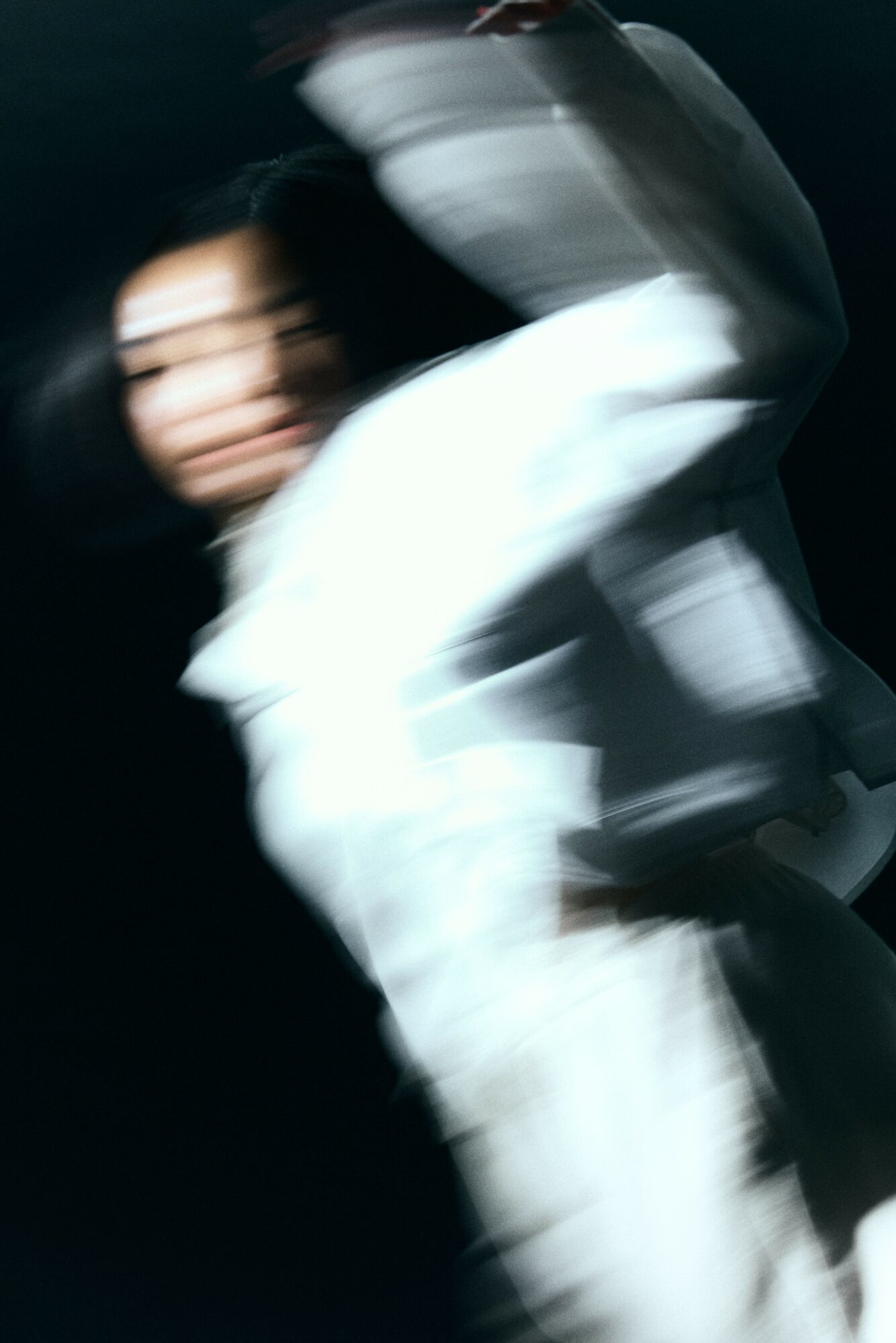 Person in white clothing with blurred motion, dark background, face partially visible, arms raised.