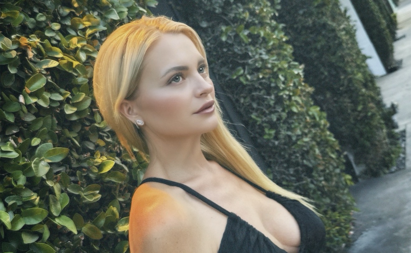Woman with long blonde hair standing outdoors near green bushes, looking to the side, wearing a black top.