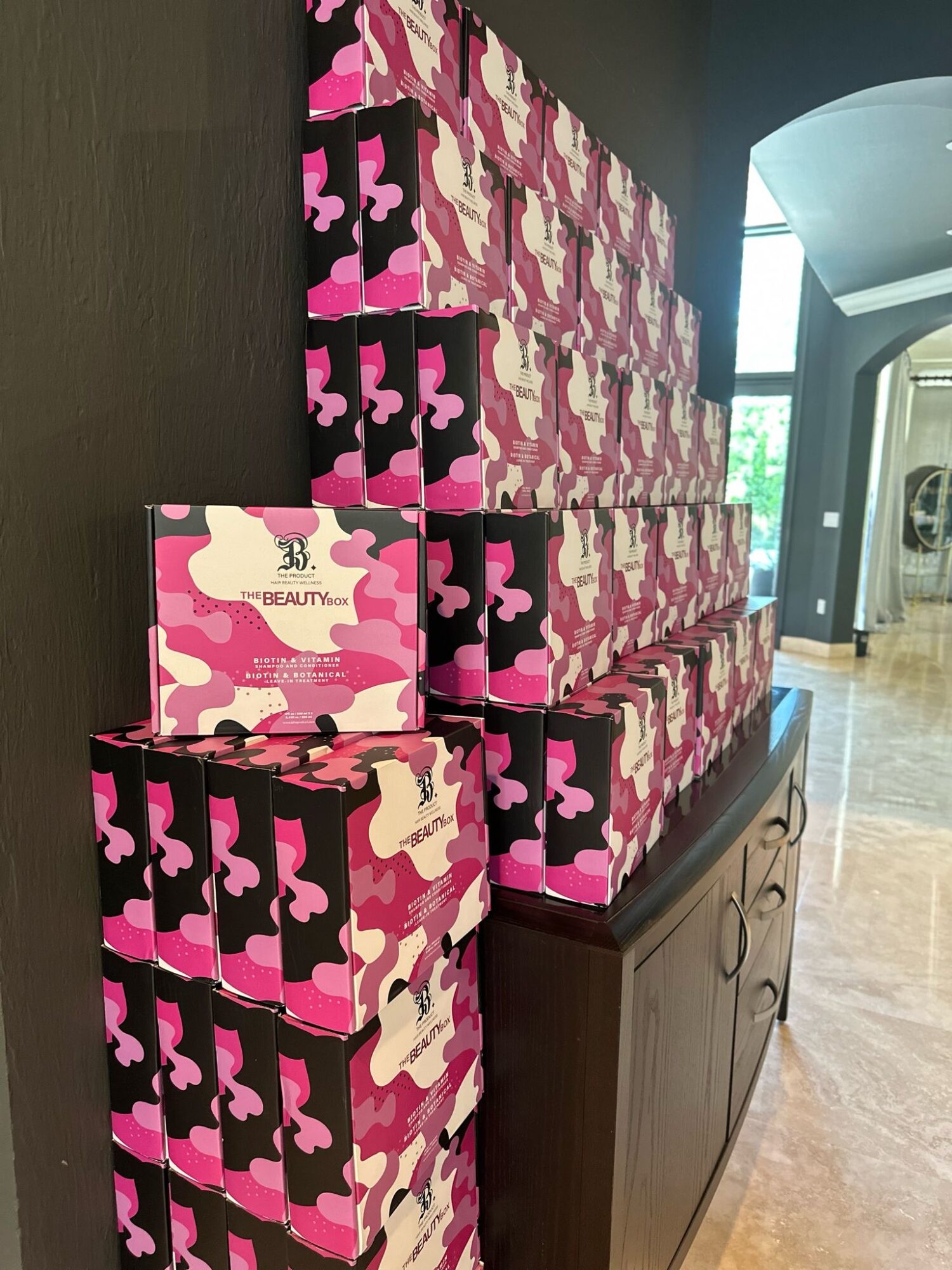 Stacked pink, black, and beige camouflage-patterned boxes on a wooden cabinet, with a sign on the front box, in a room with a window.