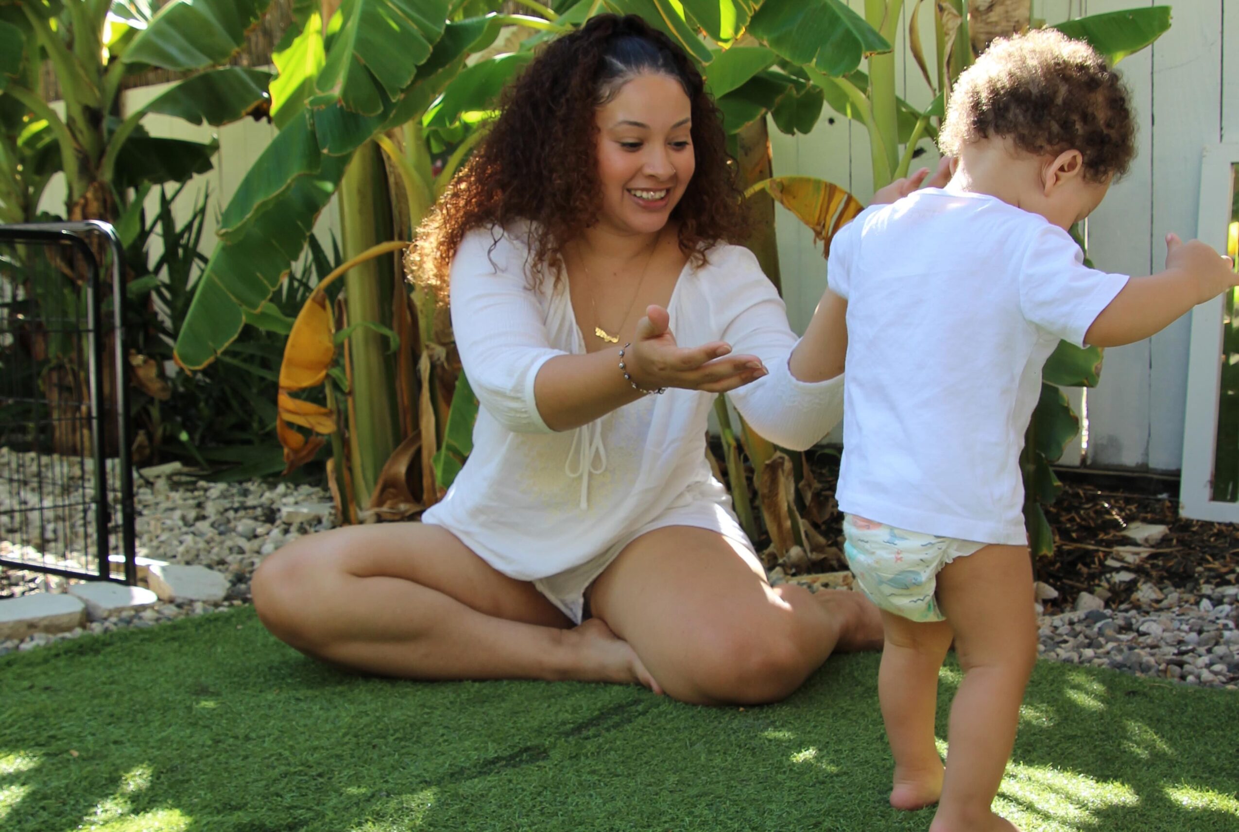 Woman sitting on grass with a child, playing outdoors near plants and a white fence.