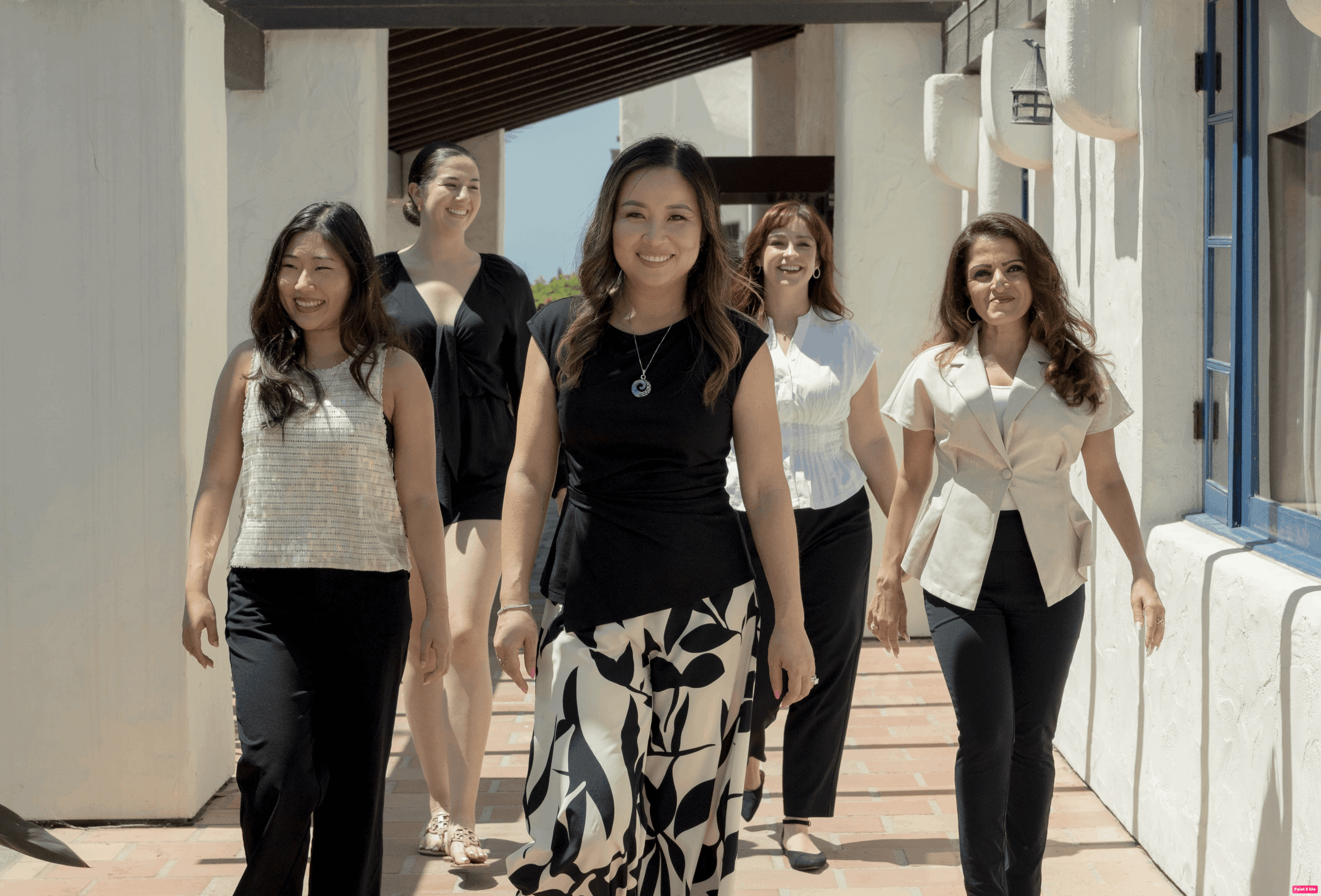 Five women walking together in a hallway, smiling, with sunlight coming through windows on the right.