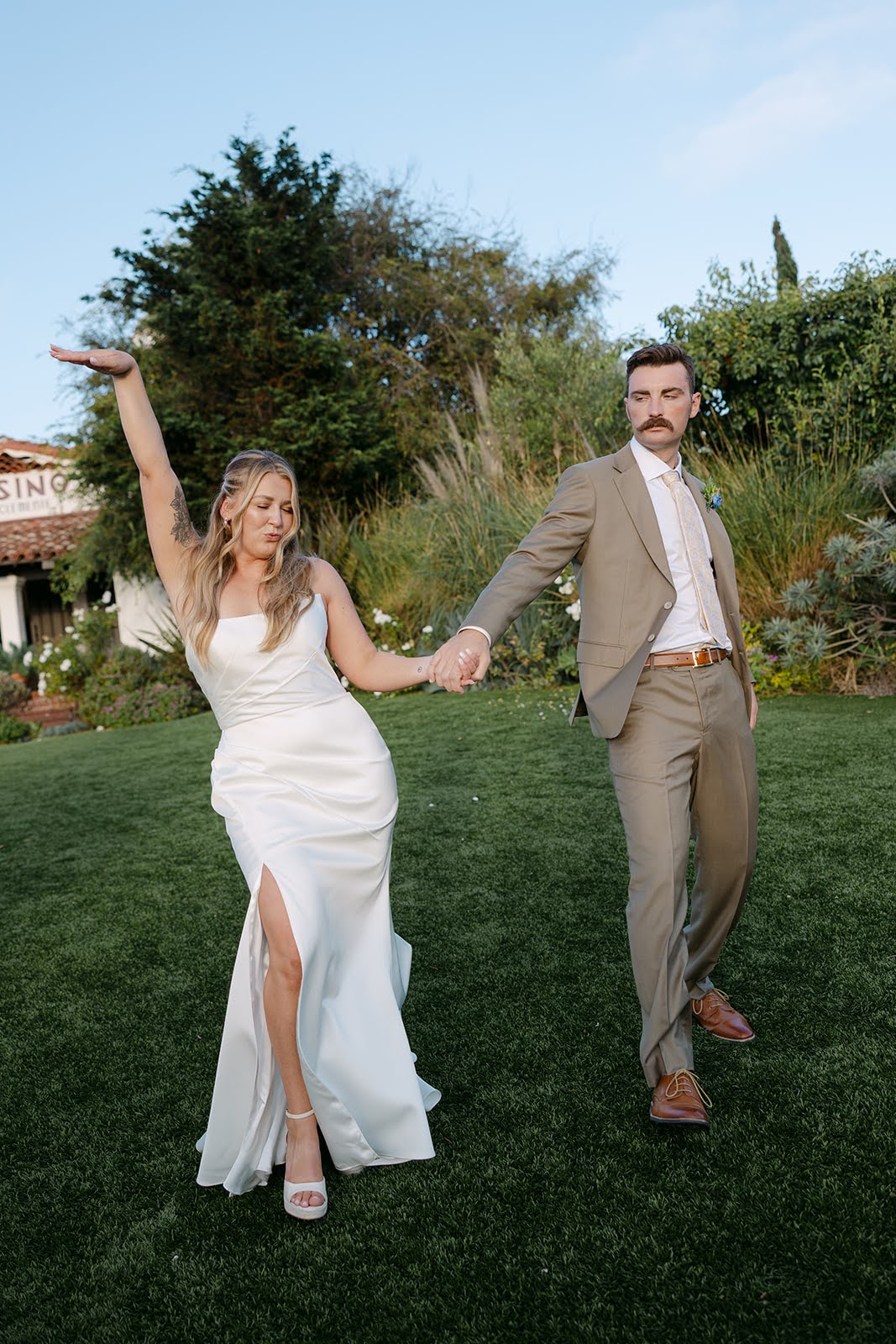A woman in a white dress and a man in a suit hold hands outdoors on grass, with trees and a building in background.