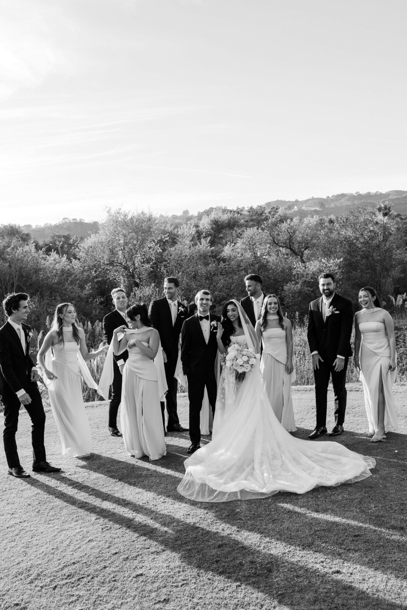 Group of people in formal attire outdoors, with a bride in a wedding gown, holding hands and standing on grass, trees in background