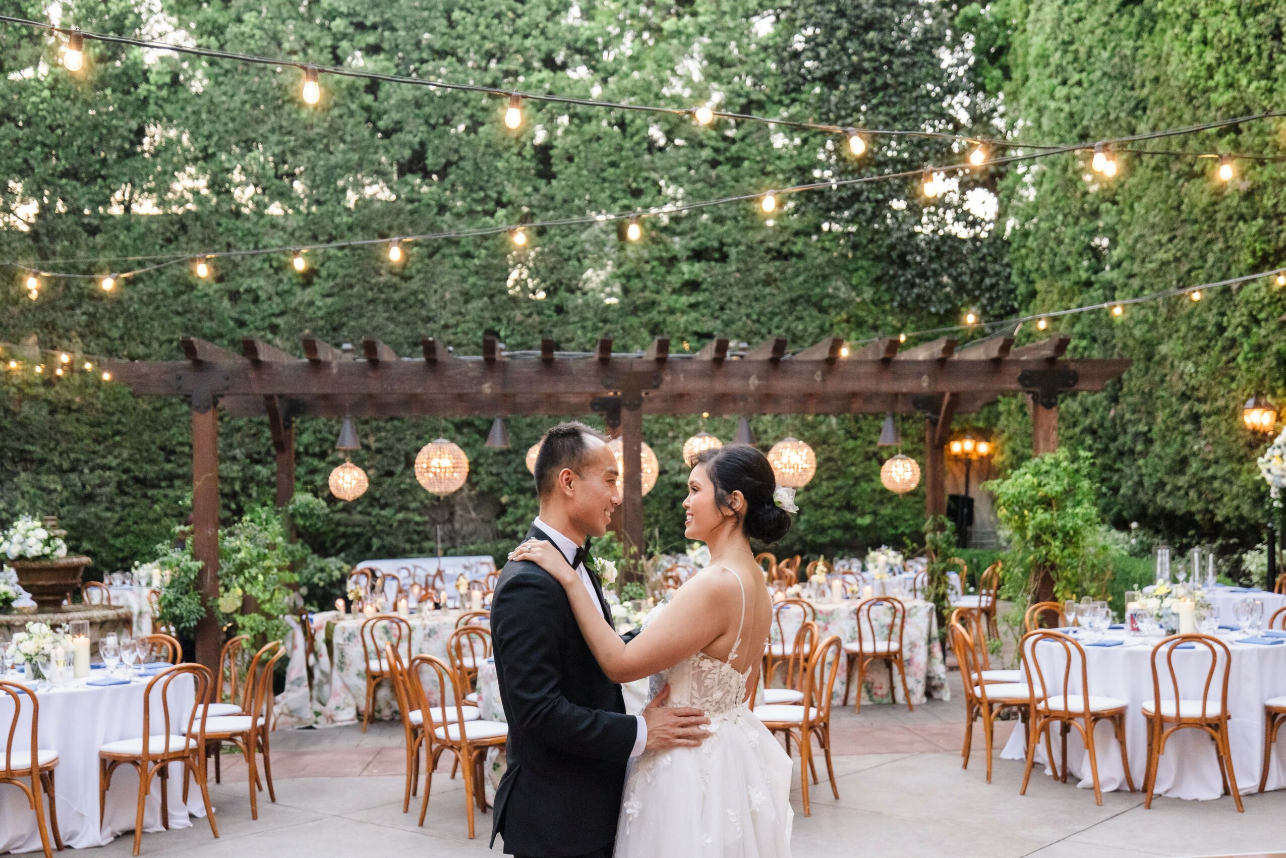 A bride and groom hold each other in an outdoor wedding reception with string lights and decorated tables.