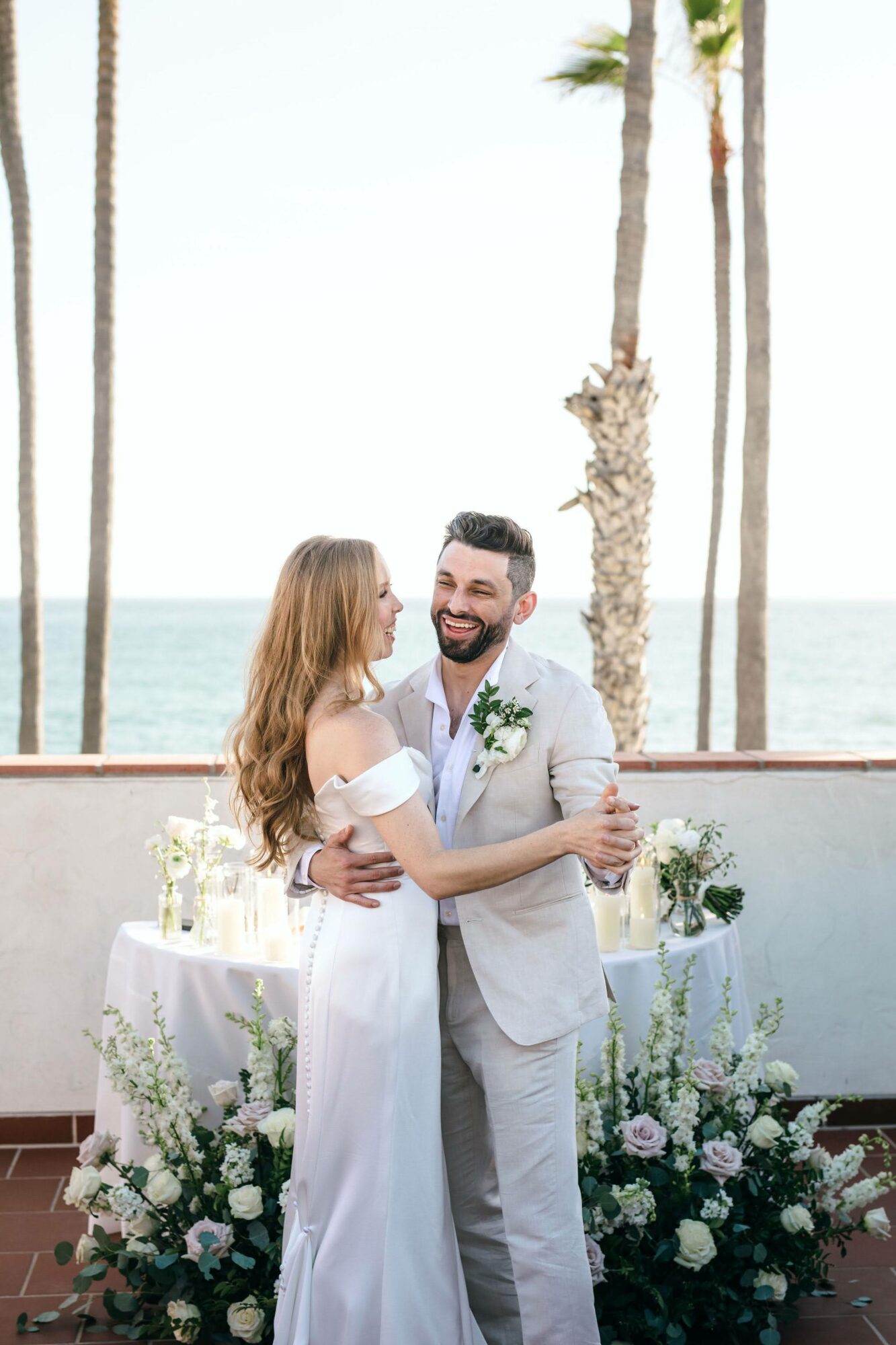 A couple in wedding attire hugging outdoors with ocean, palm trees, and a decorated table behind them.