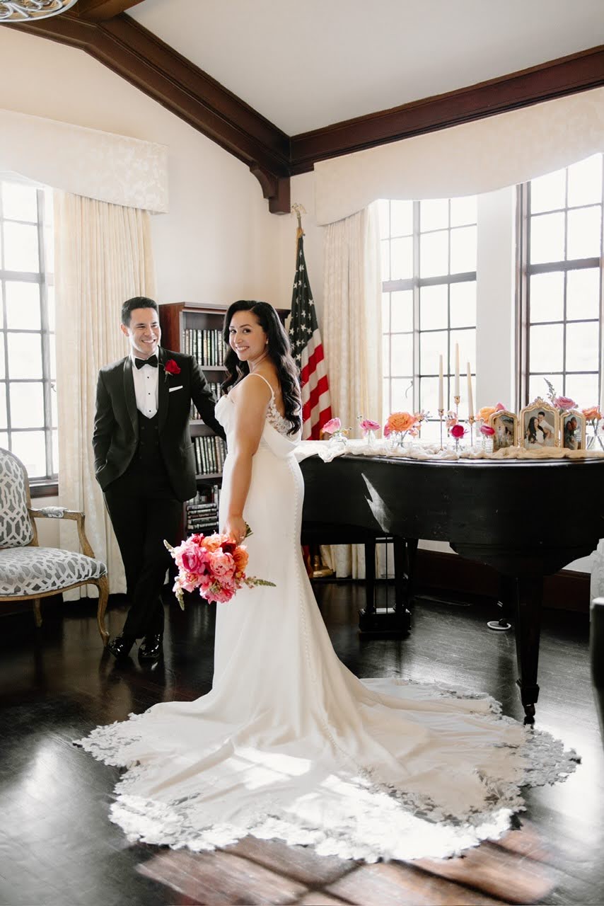 Bride in a white wedding dress holding a bouquet, groom in a tuxedo, standing in a decorated room with a grand piano and American flag.