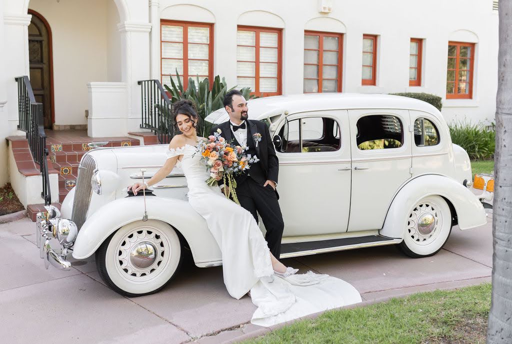 Bride and groom sit on a vintage white car in front of a house, bride holding a bouquet, groom in a suit.