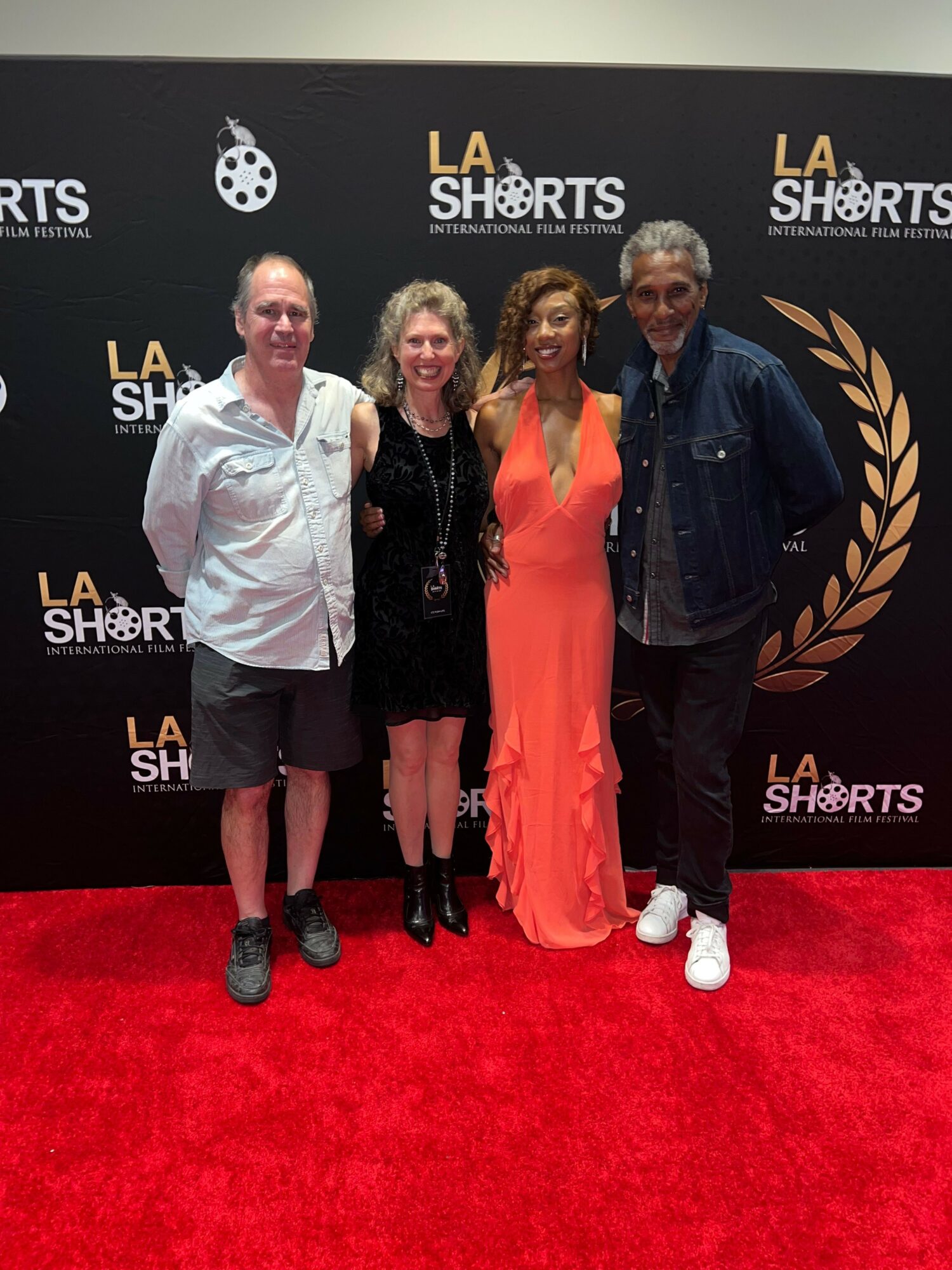 Four people standing on a red carpet in front of a black backdrop with gold laurel and text, posing for a photo.