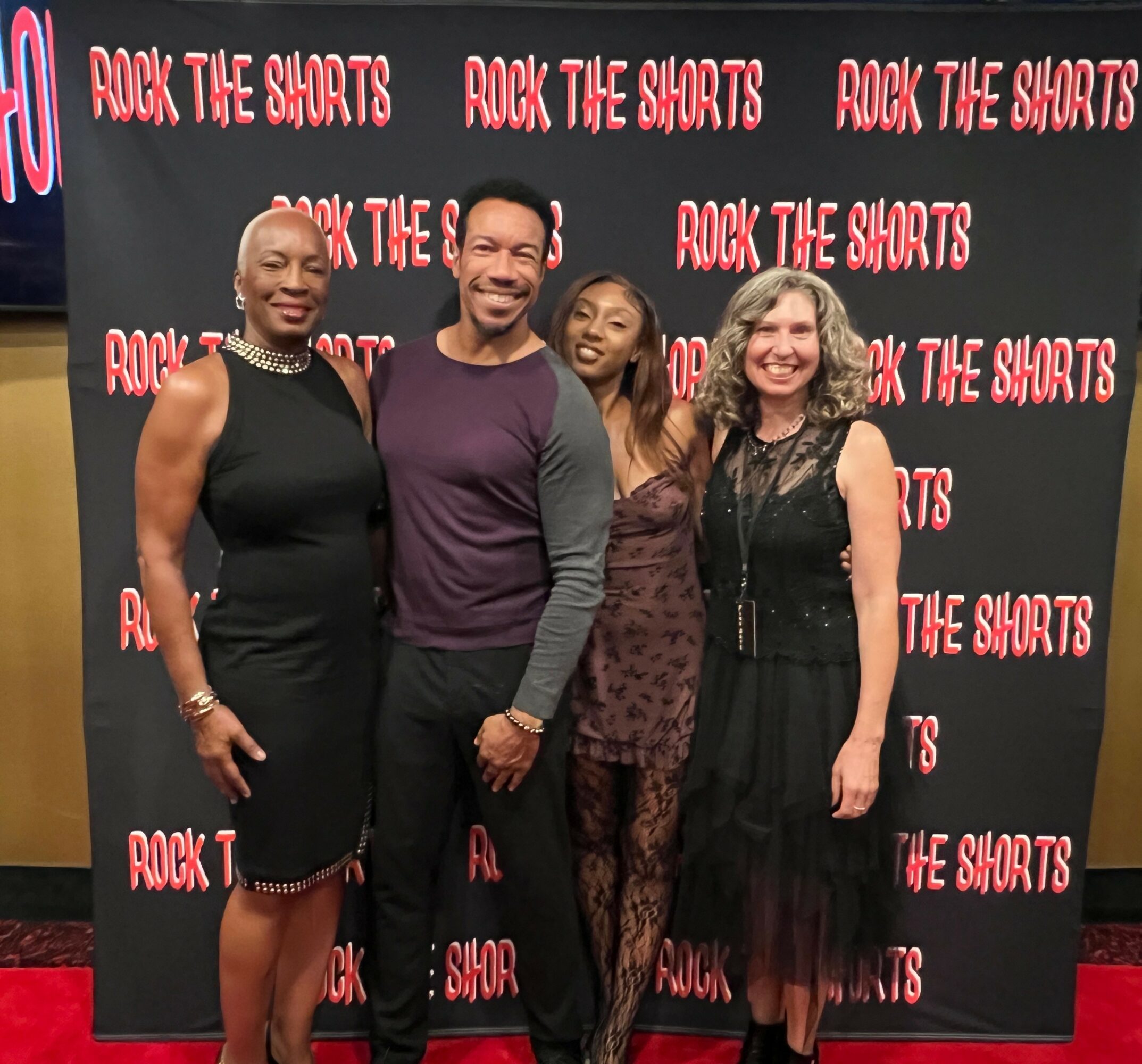 Four people standing in front of a backdrop with red and pink text, smiling at the camera.