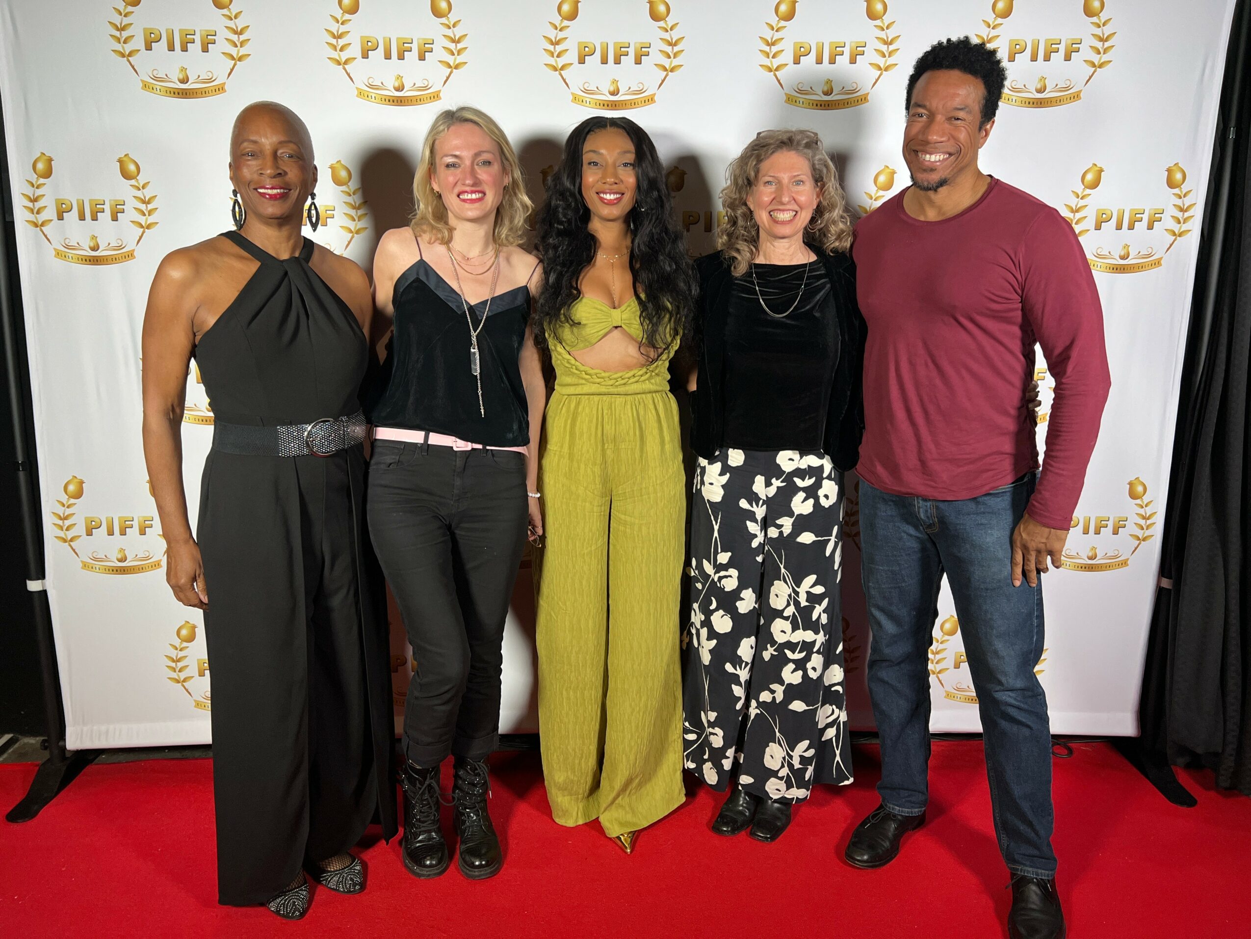 Five people standing on a red carpet in front of a white backdrop with gold logos, smiling and posing for a photo.