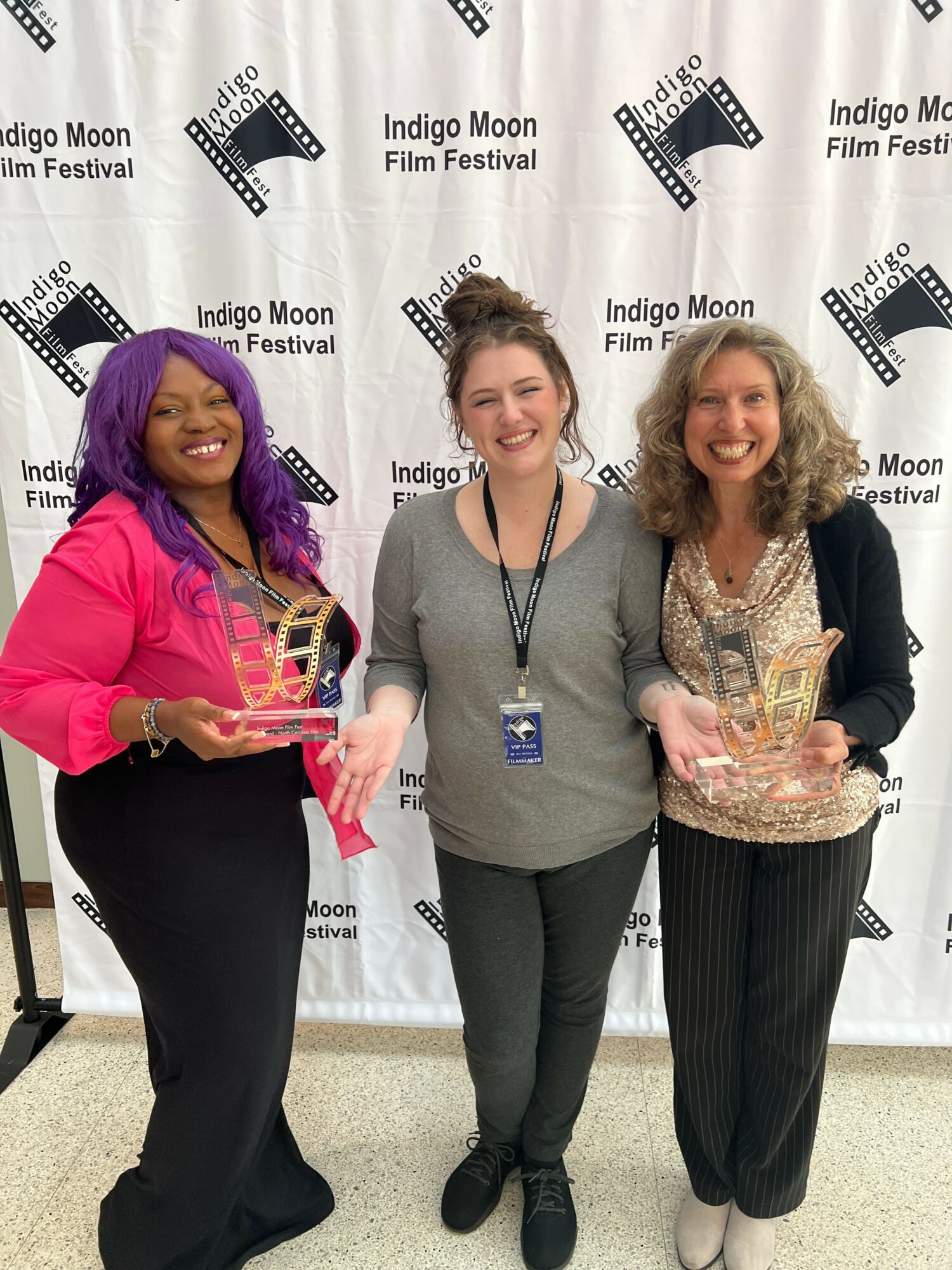 Three women standing together, one holding an award, smiling, in front of a backdrop with film reel graphics and text.