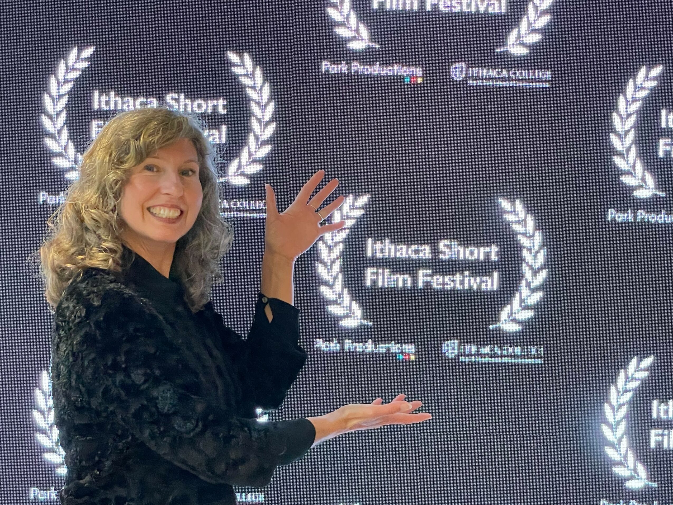 Woman with curly hair smiling and gesturing in front of a backdrop with festival logos.