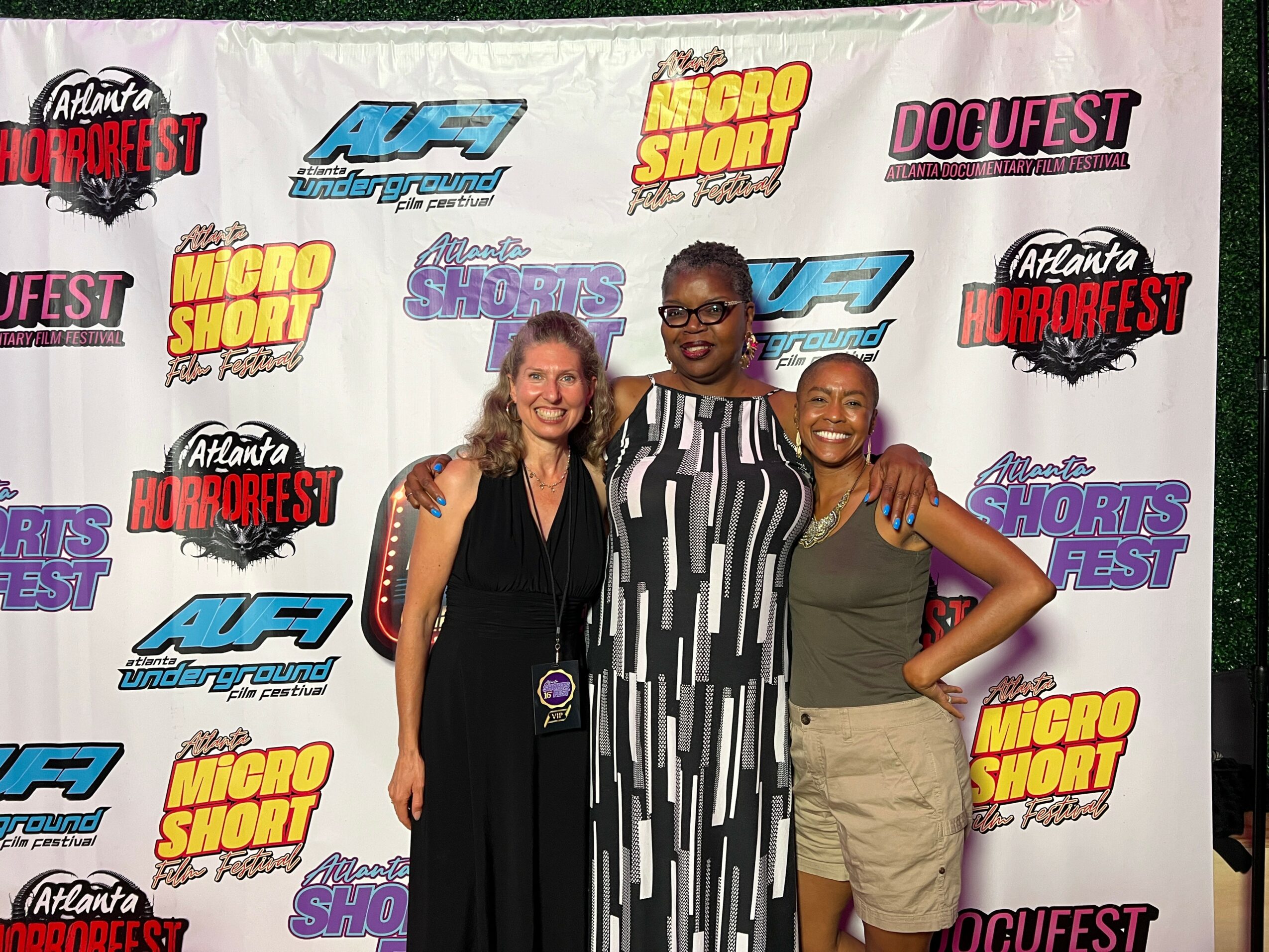 Three women standing in front of a backdrop with event logos, smiling, with one woman wearing glasses and a striped dress.