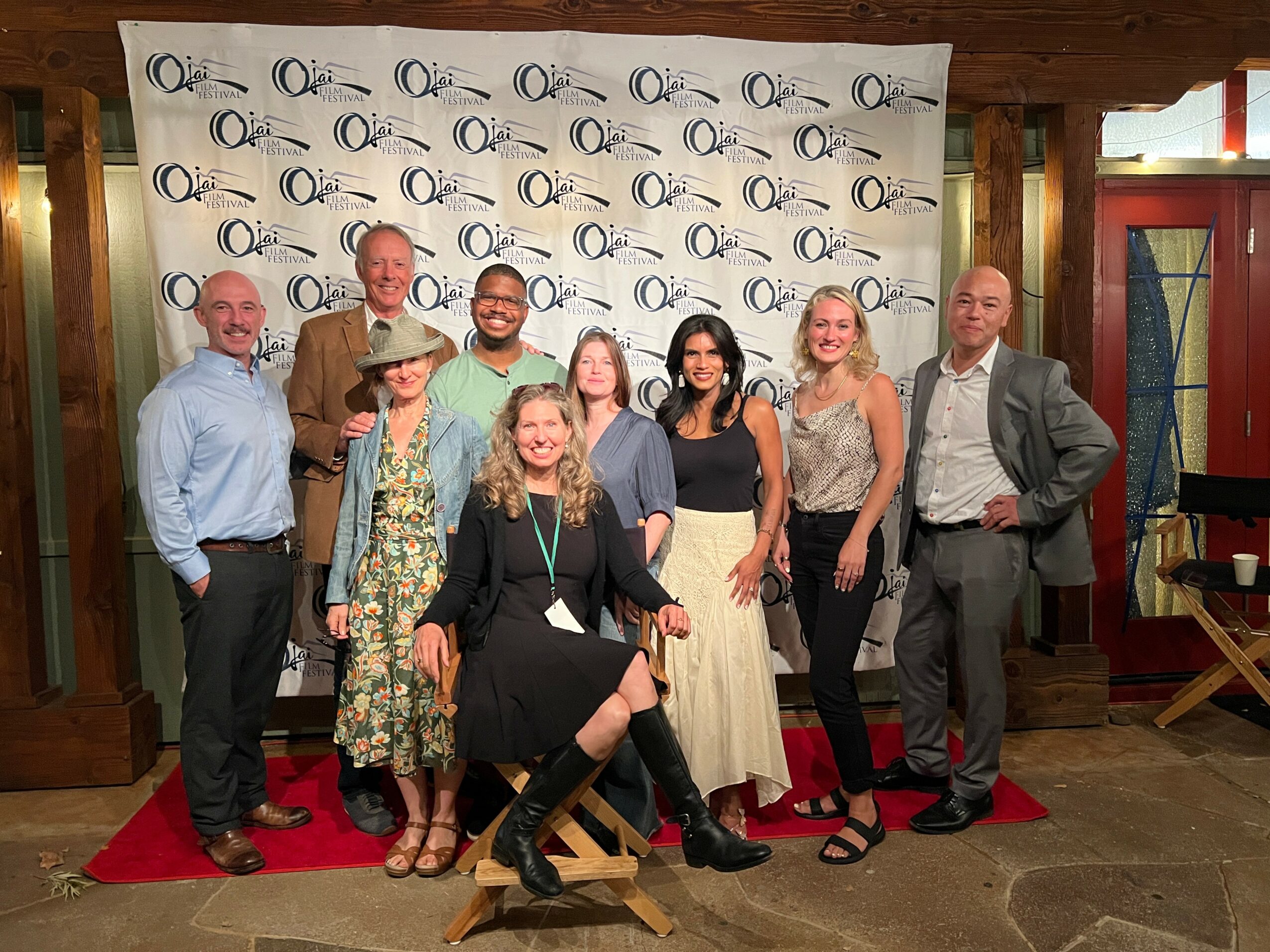 Group of ten people standing on a red carpet in front of a backdrop with a logo, smiling for the photo.