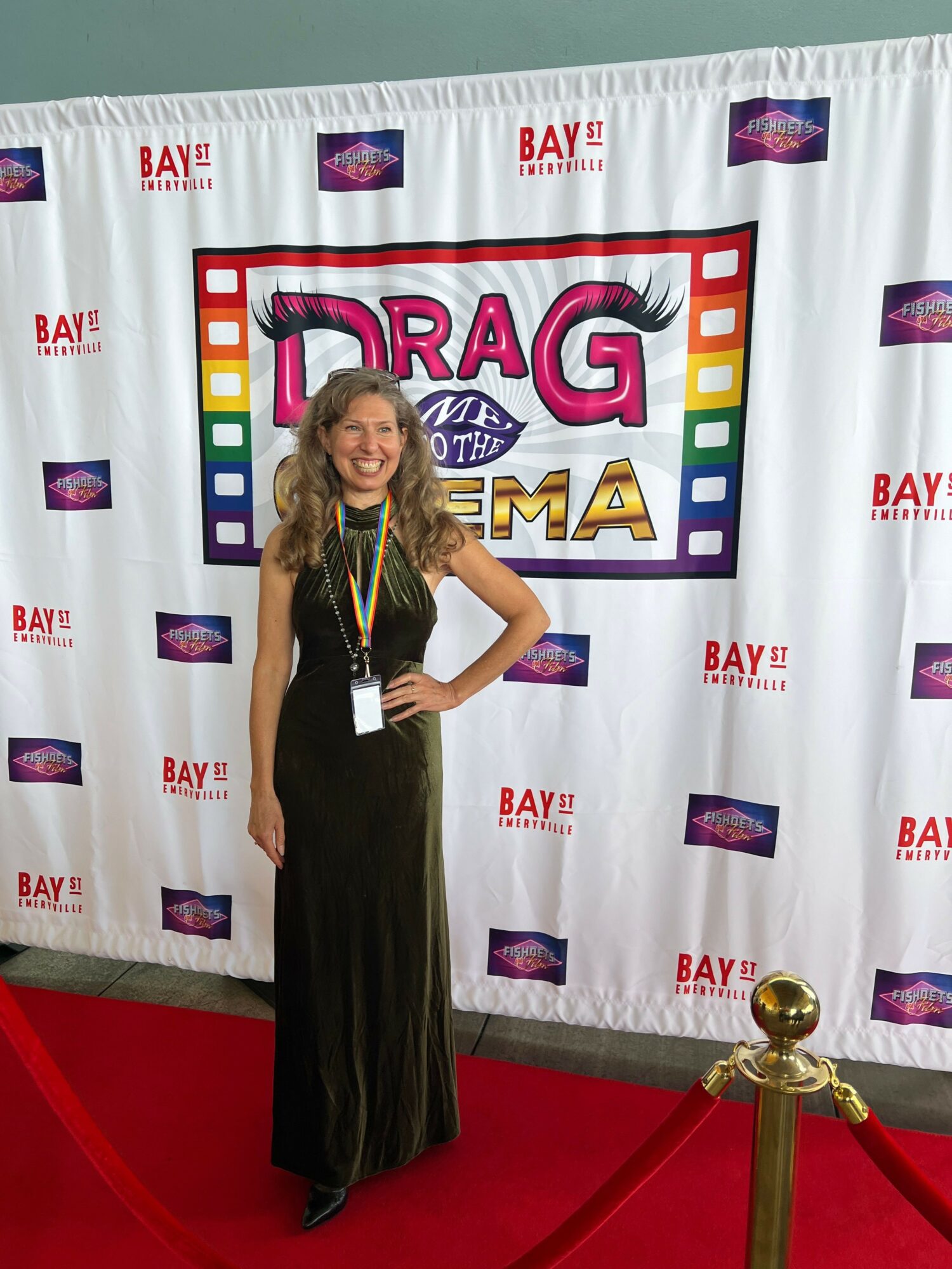 Woman in black dress standing on red carpet in front of a backdrop with colorful logo and text, holding a phone.