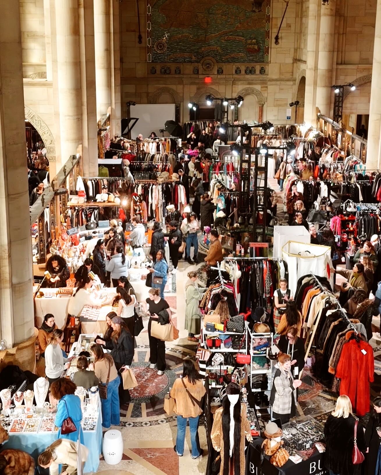 Indoor market with stalls and shoppers browsing clothing and accessories, high ceiling with columns, busy atmosphere.