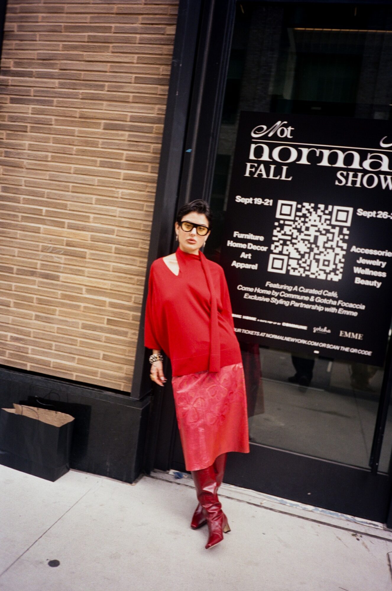 Woman in red outfit and glasses standing outside near a glass door with a poster, brick wall background.