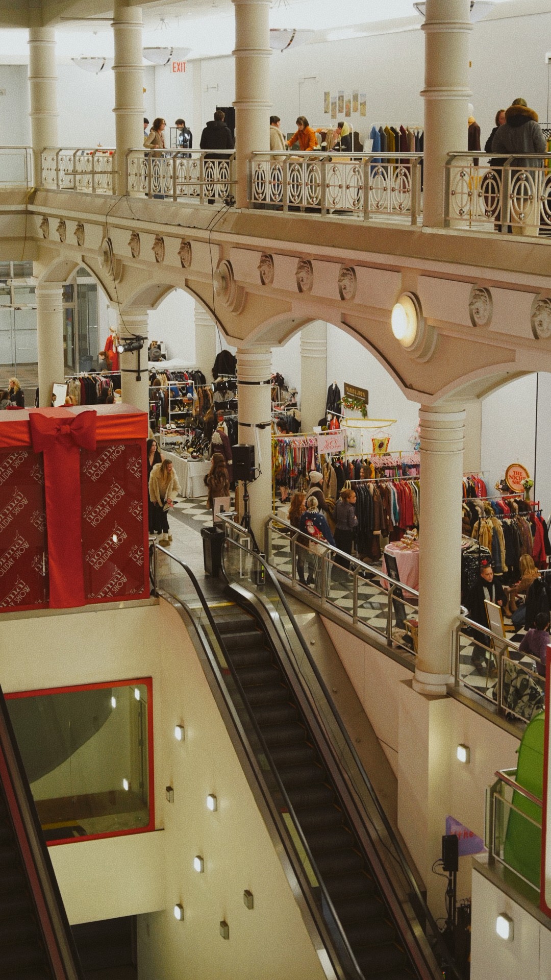 People shopping and walking on upper and lower levels of a shopping mall with arches and columns.