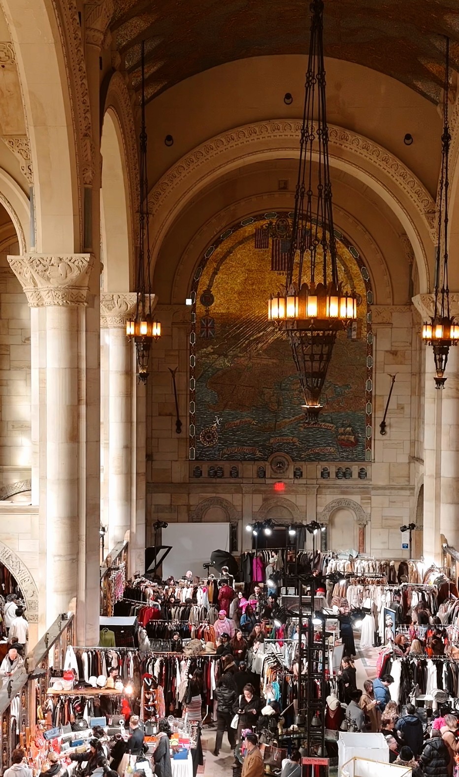 Interior of a large hall with high arched ceilings, chandeliers, and a busy market with stalls and people.