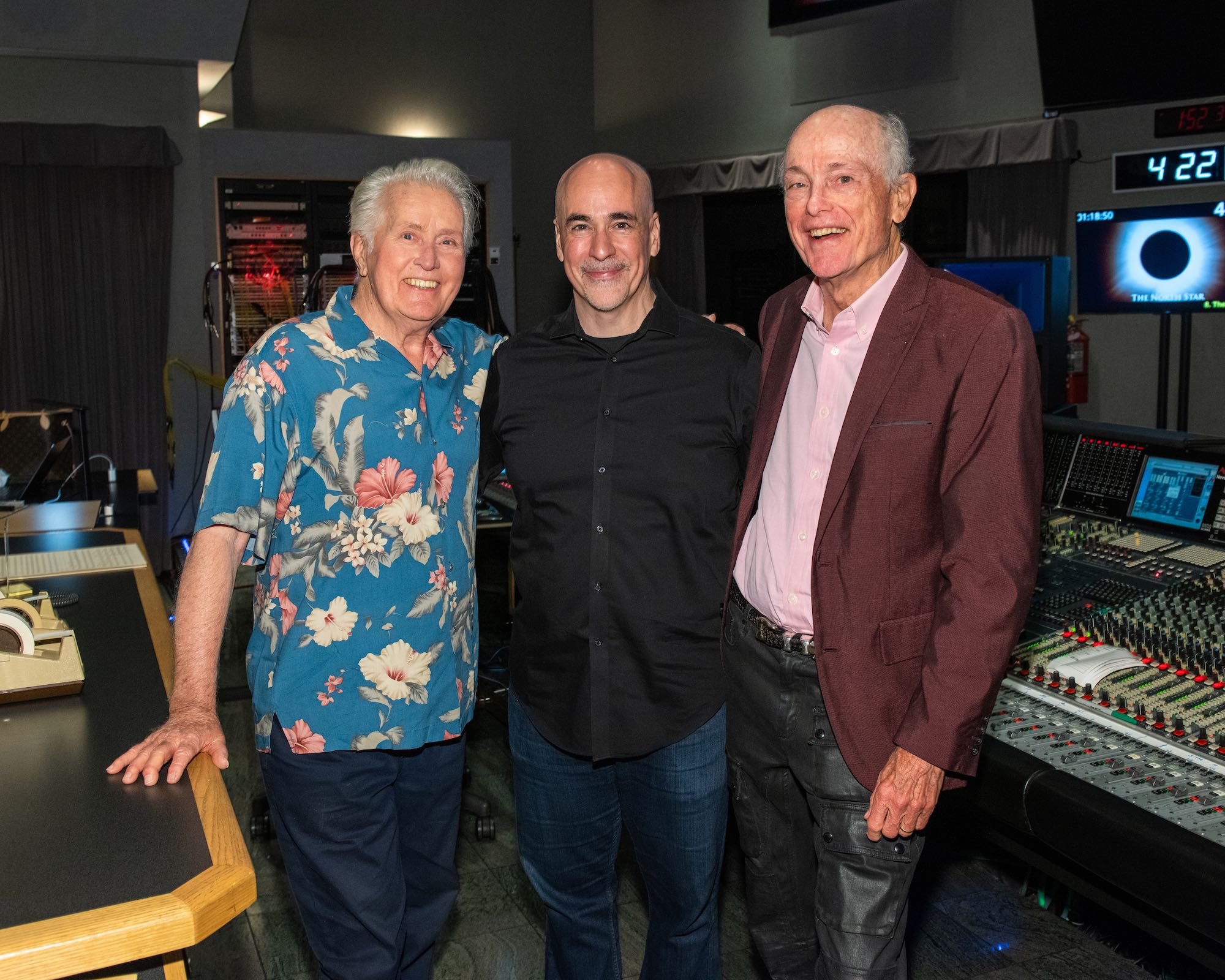 Three people standing together in a room with audio equipment, smiling at the camera.