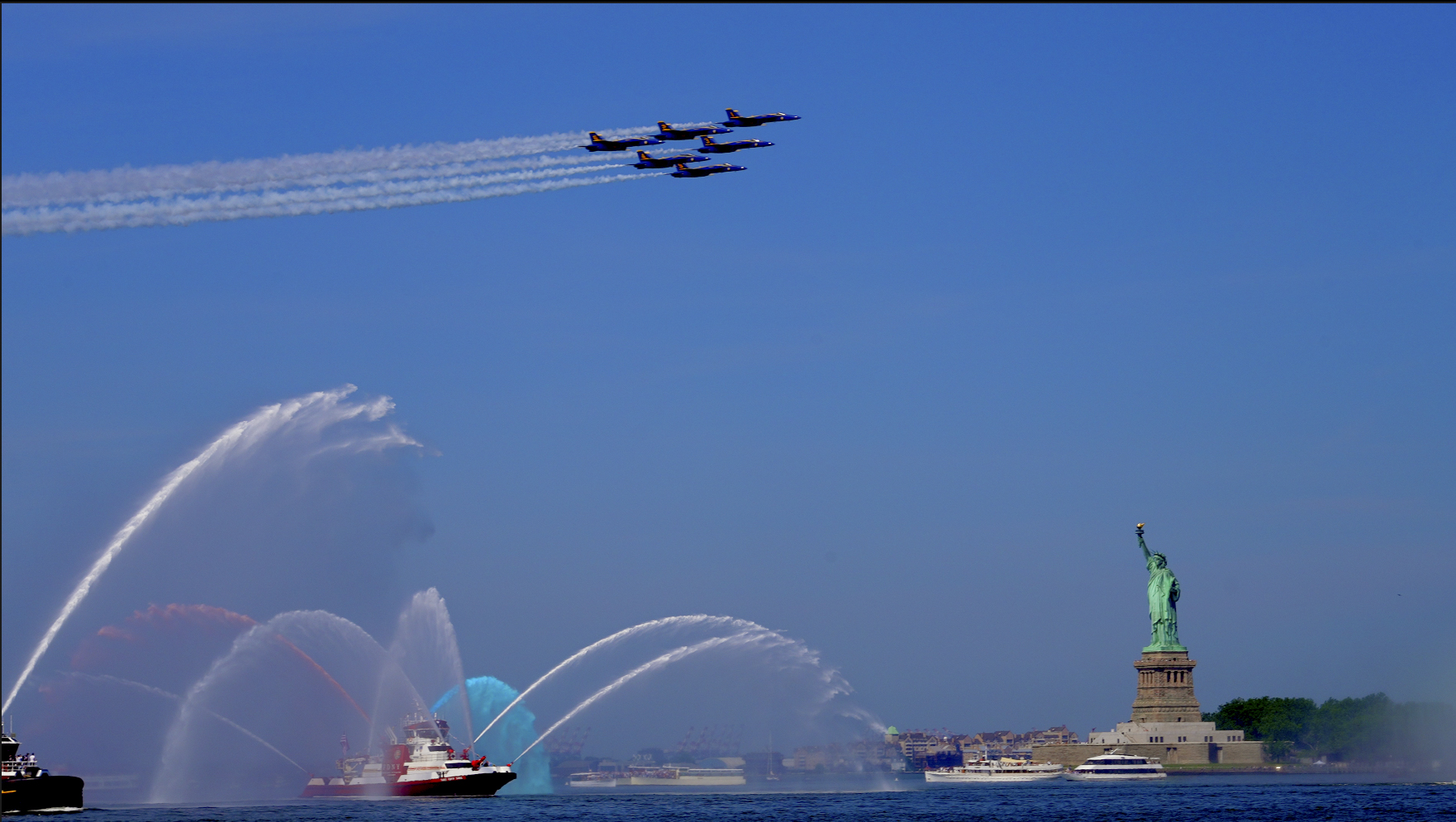 Four jets fly in formation above water, leaving trails, with the Statue of Liberty and water fountains in the background.
