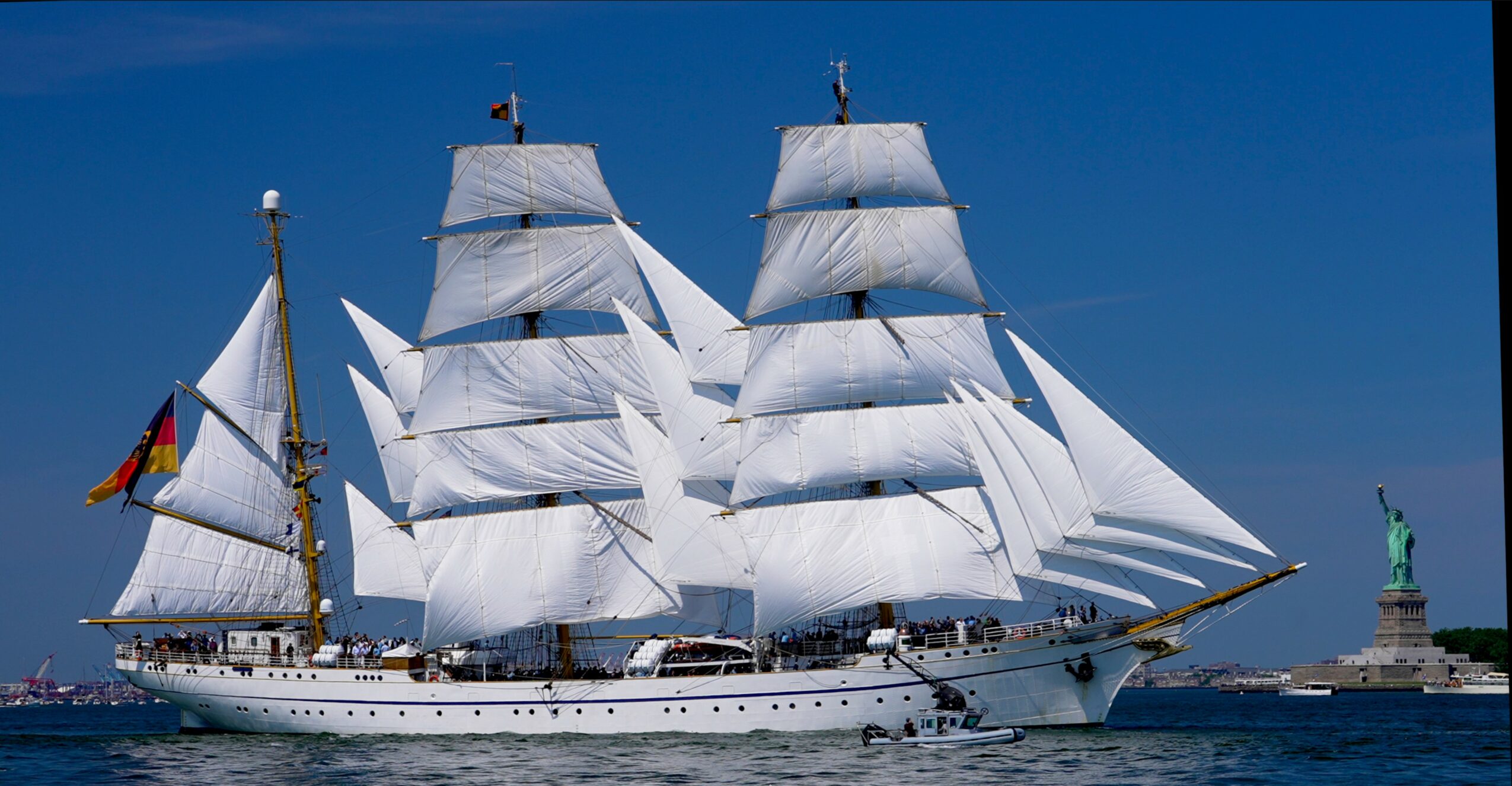Large sailing ship with white sails on the water, with a lighthouse in the background.