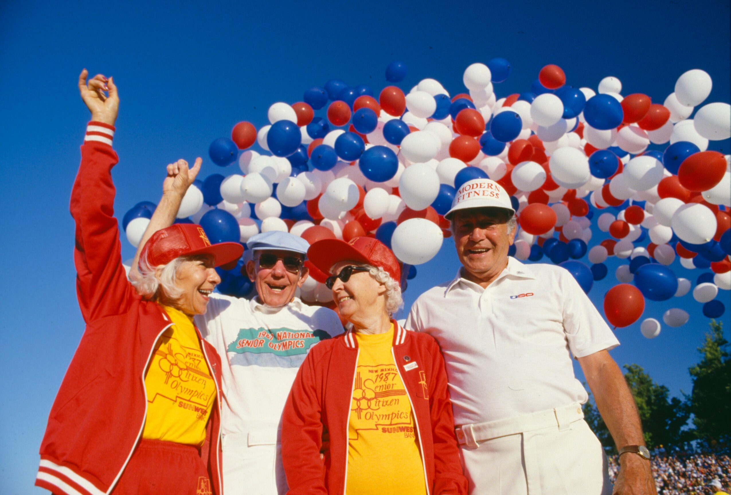 Four people in colorful clothing and hats release red, white, and blue balloons into the sky during a celebration.