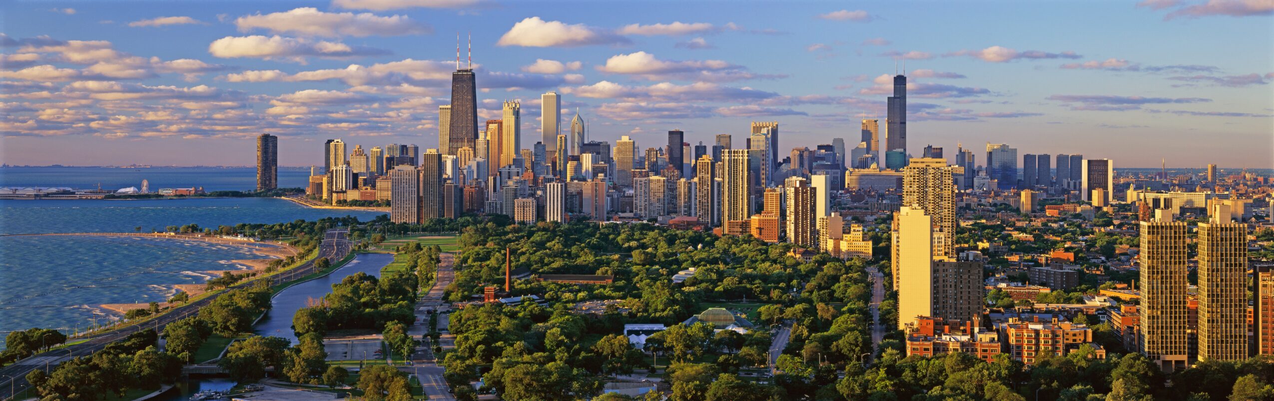 City skyline with tall buildings, a river, and green parks under a partly cloudy sky.