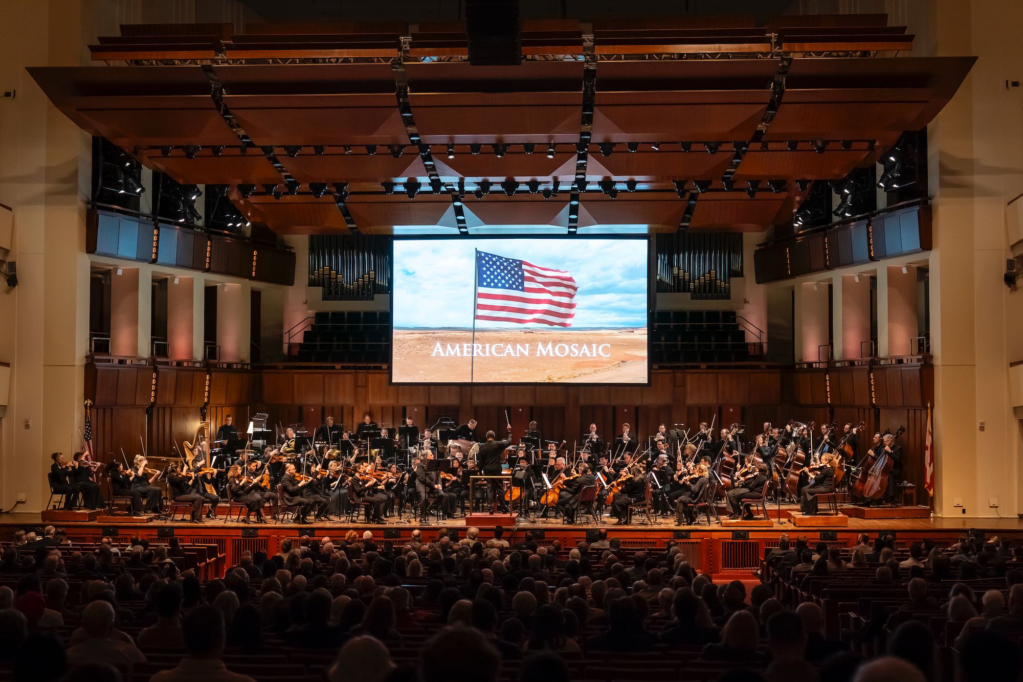 Orchestra performing on stage with a large screen displaying an American flag and text, audience watching, concert hall setting.