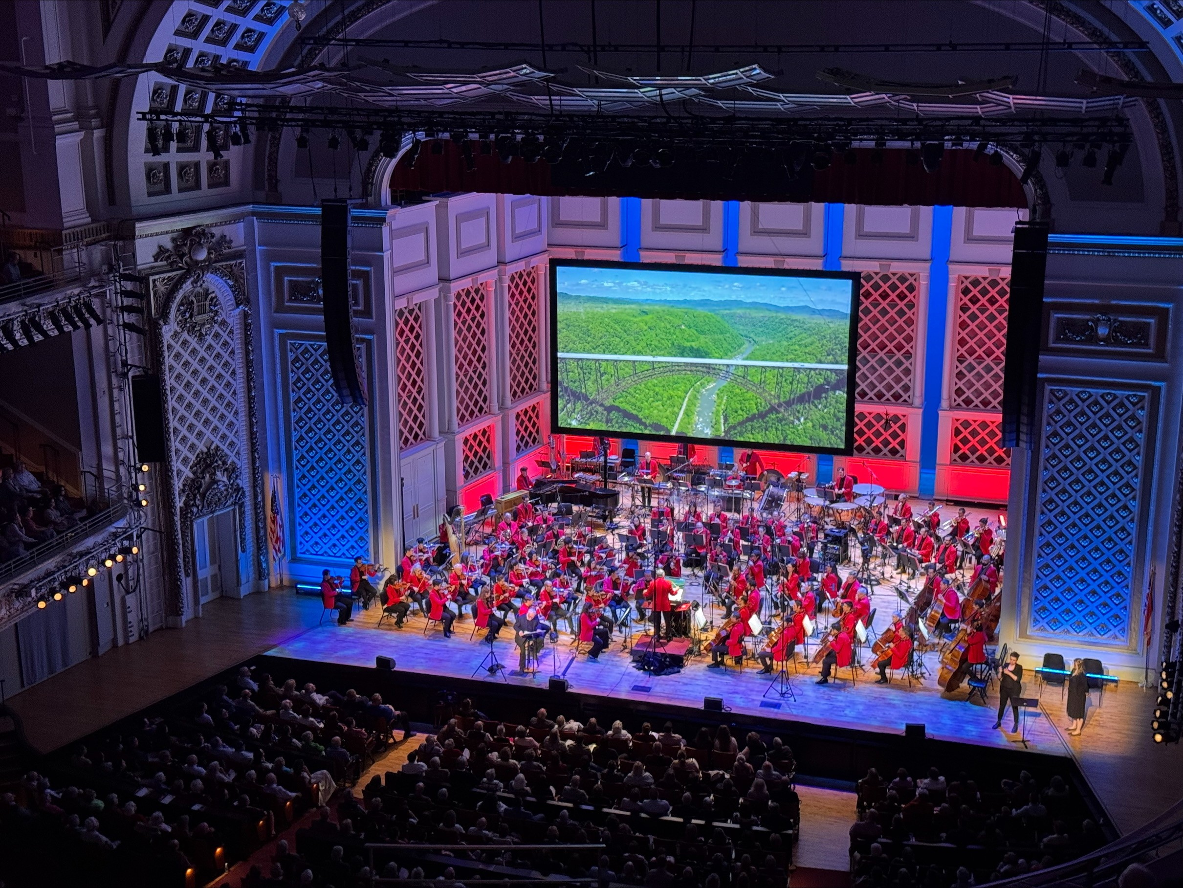 Orchestra performing on stage with a large screen showing a green landscape, audience seated in a concert hall.
