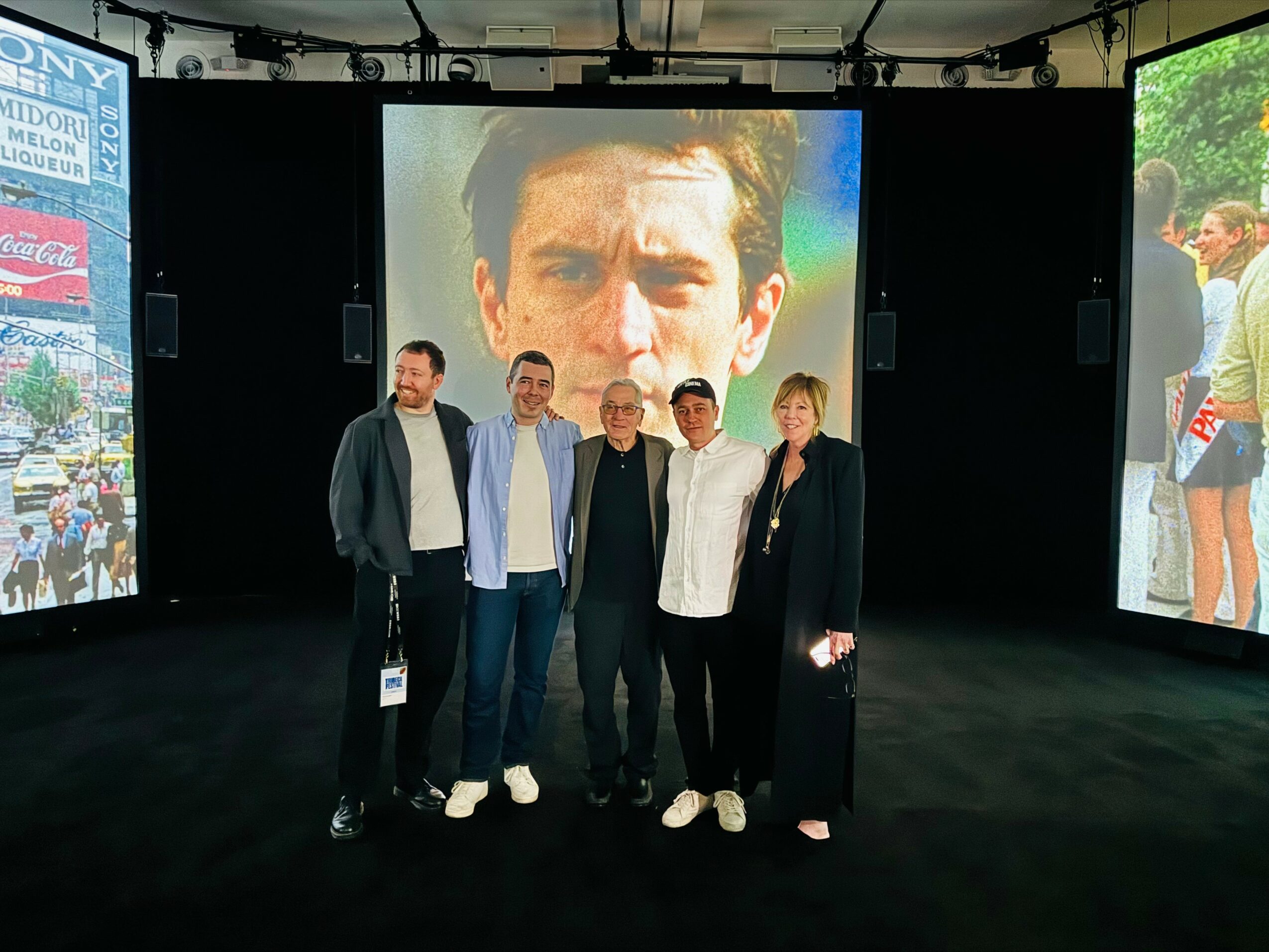 Five people standing in front of a large screen displaying a man's face, with two side screens showing outdoor scenes.