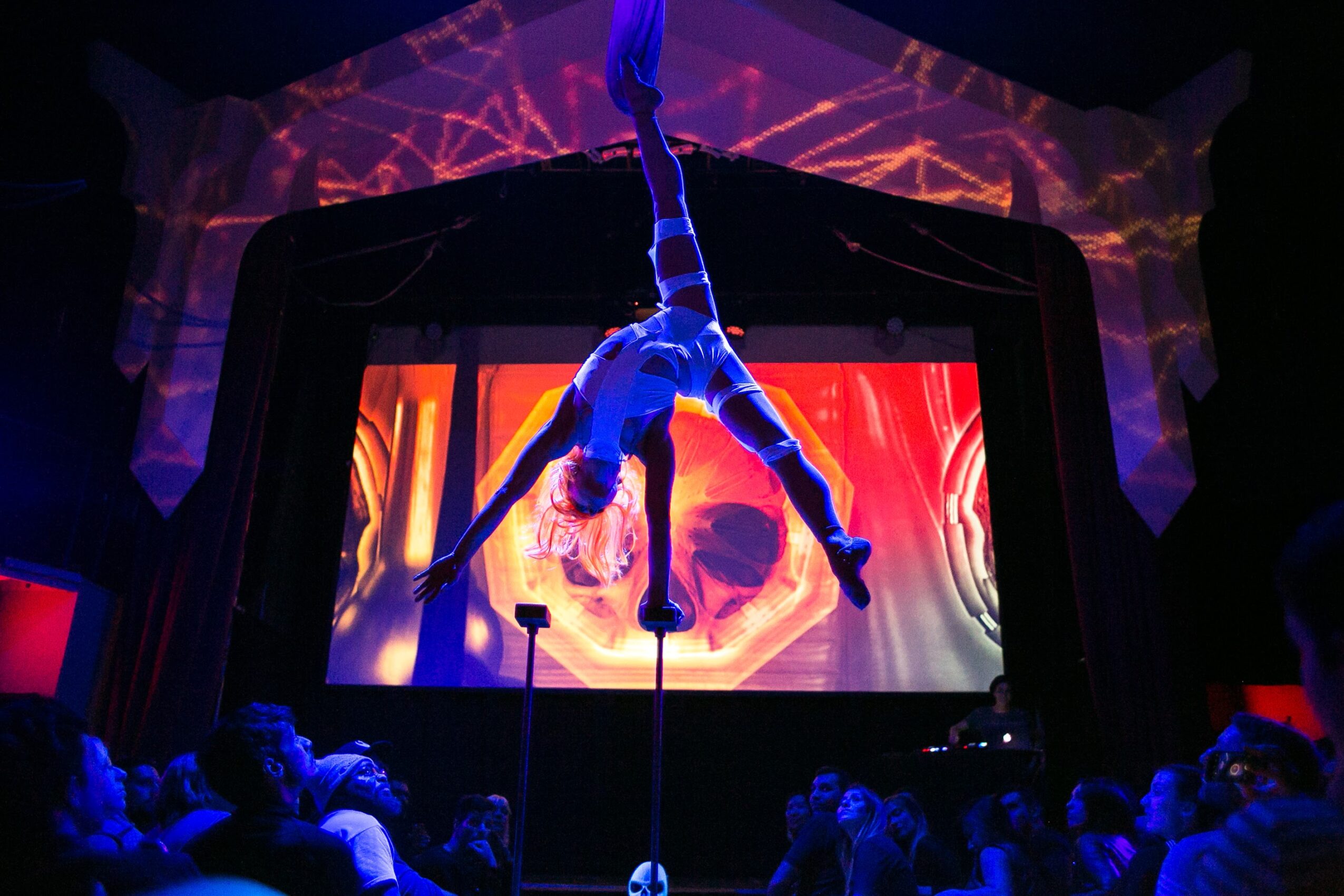 Aerial performer hangs upside down on fabric above stage with a skull projection in background, audience watching below.