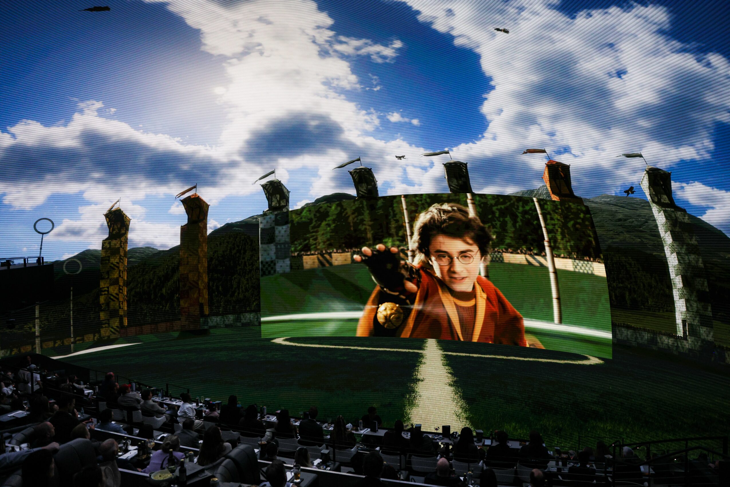 Large outdoor screen displays a person with dark hair and glasses, with a crowd watching in front, under a partly cloudy sky.