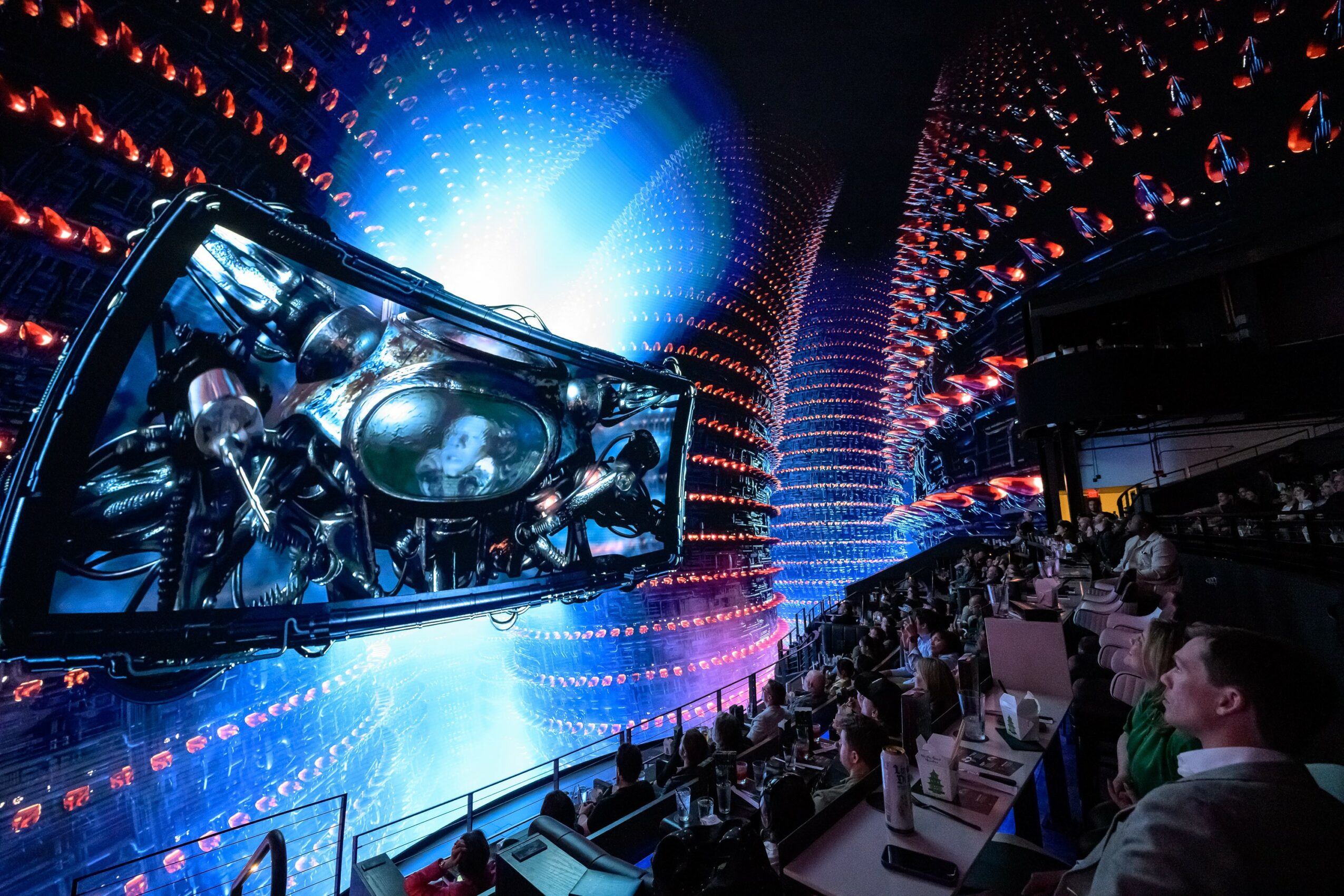 Audience watching a large screen displaying a futuristic scene with bright blue and white lights, in a dark, modern venue.