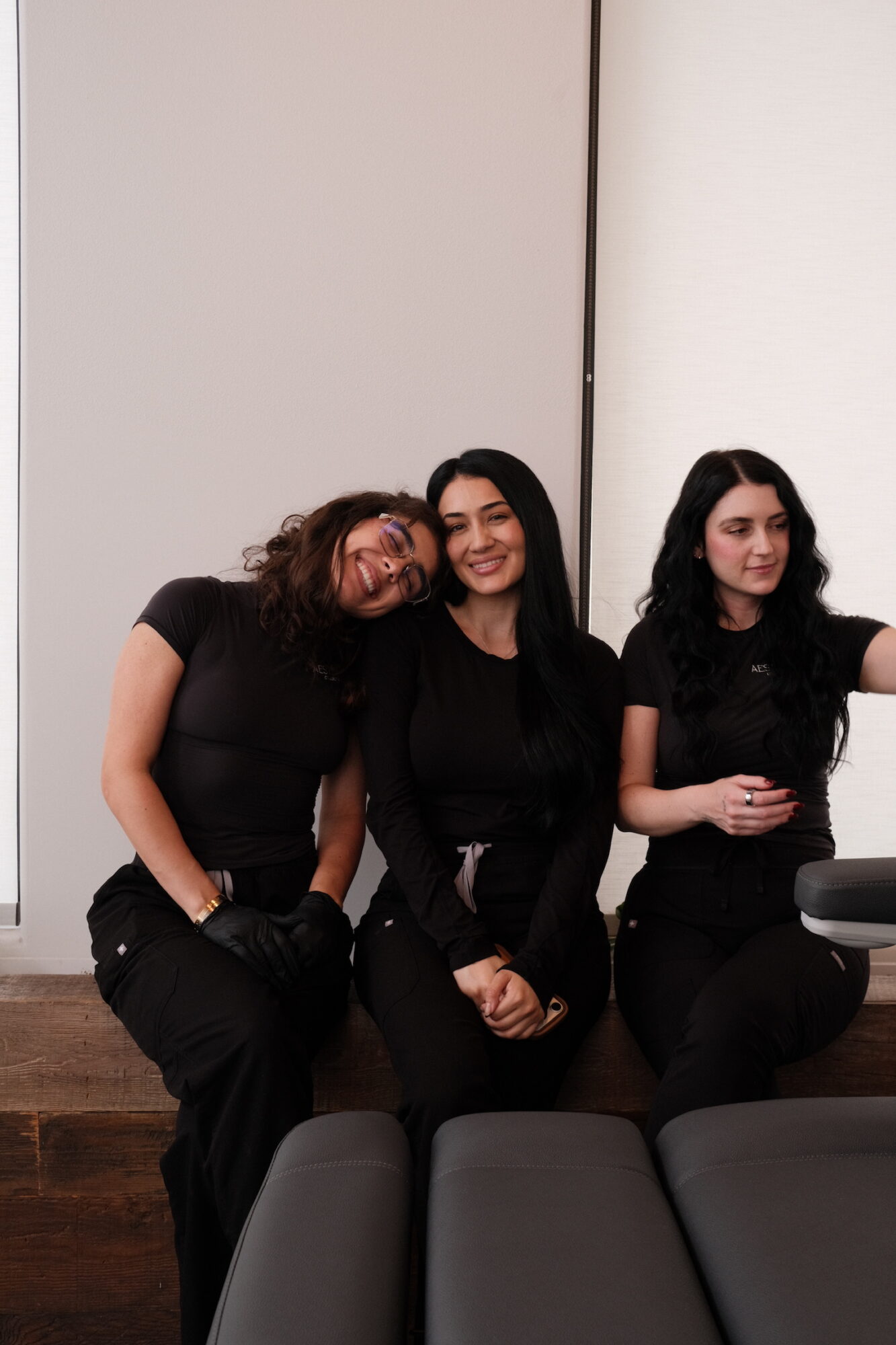 Three women sitting together, smiling, in a room with plain walls and dark flooring.