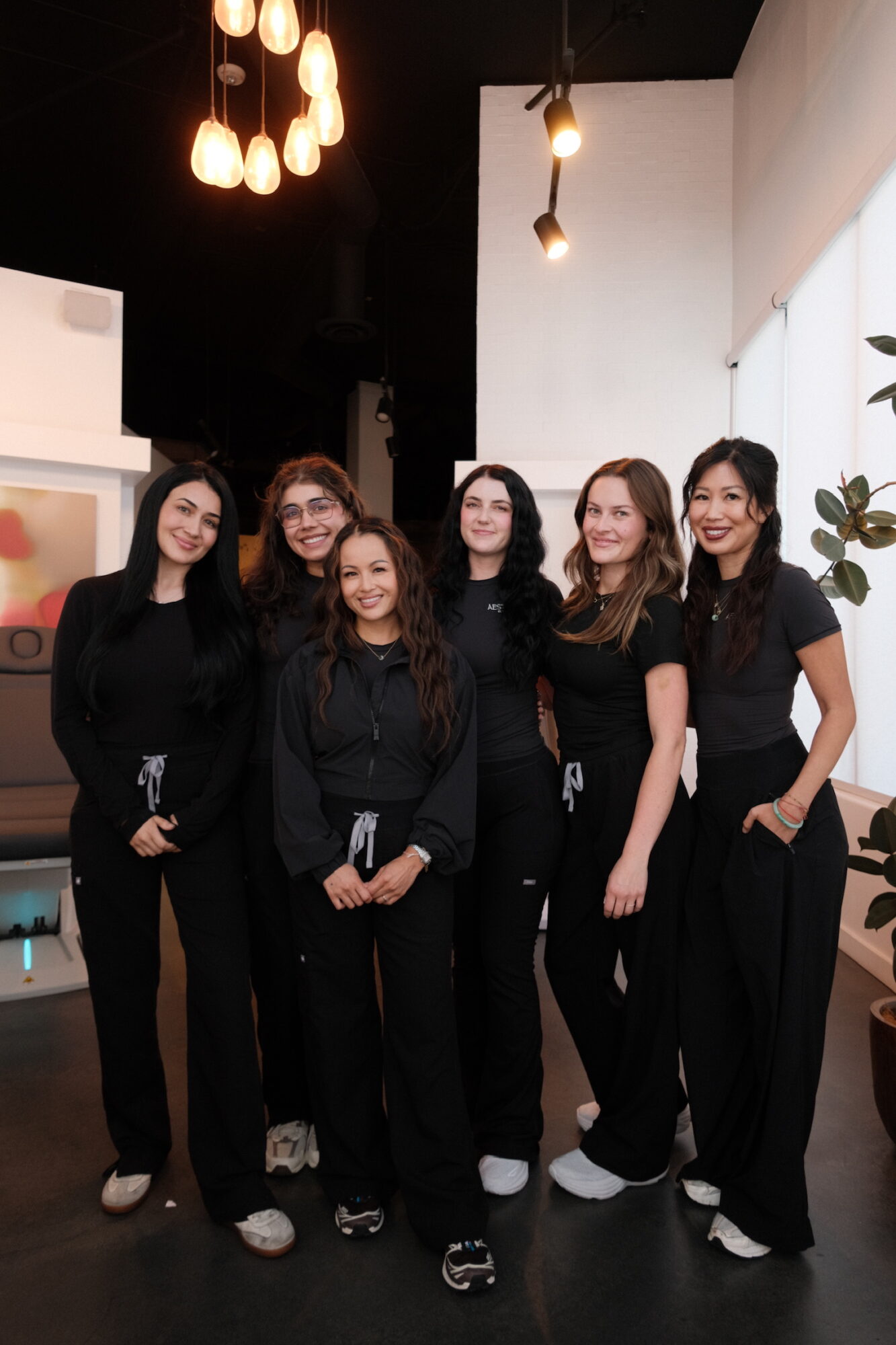 Six women in black outfits standing together indoors, smiling, with modern lighting and decor in the background.