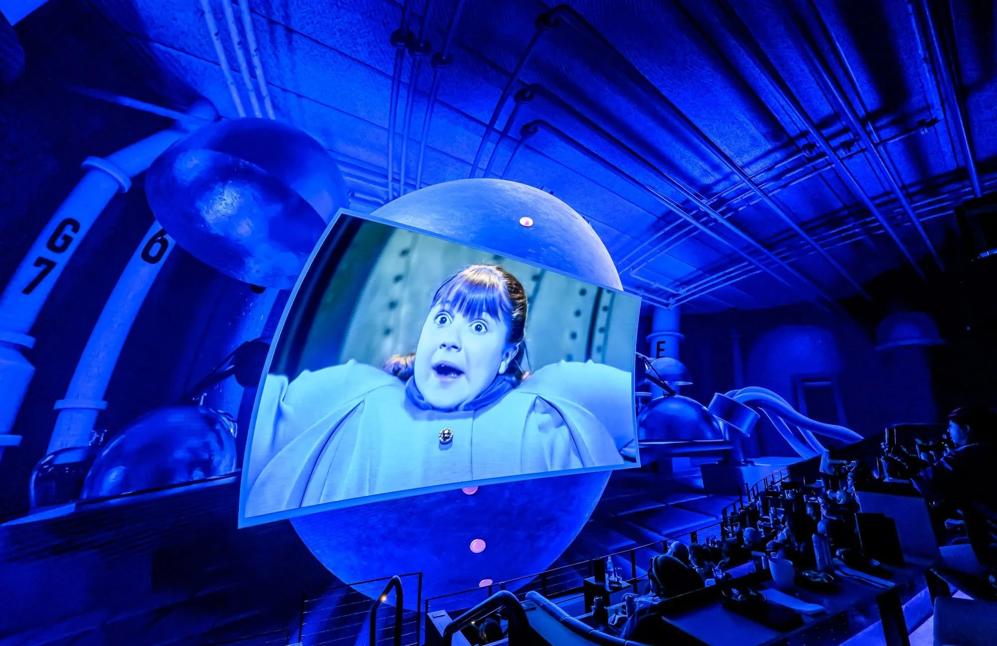 A person with a surprised expression appears on a large screen in a futuristic, blue-lit environment.