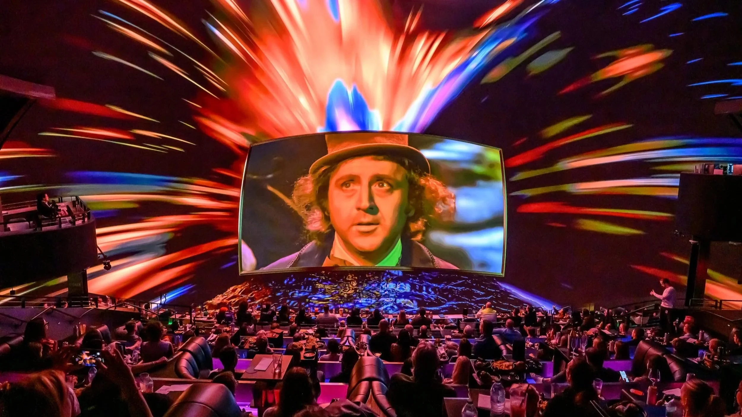 A large screen shows a woman with curly hair, surrounded by colorful light streaks, in a theater with an audience.