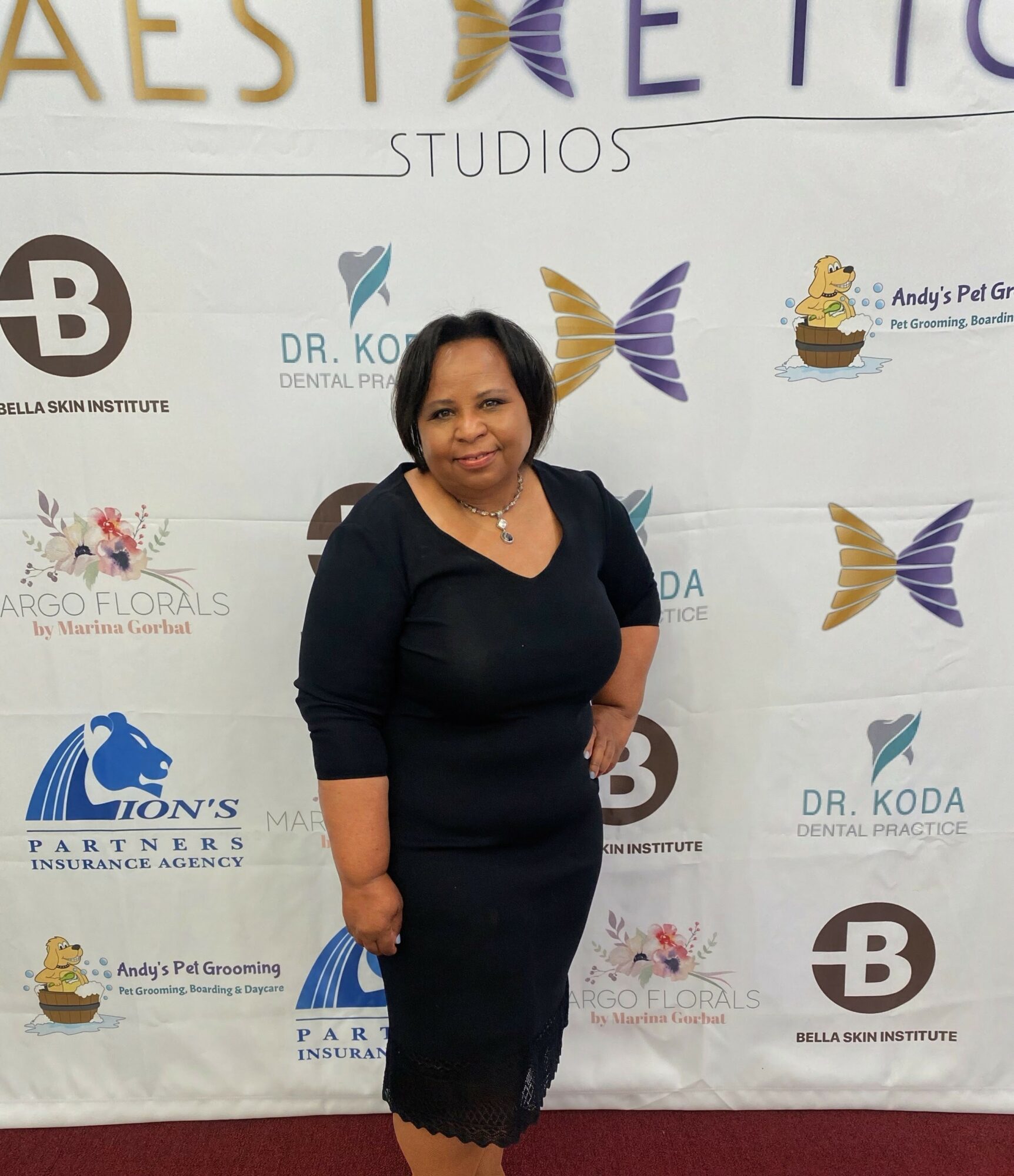 Woman in black dress standing in front of a step-and-repeat banner with various logos and text.
