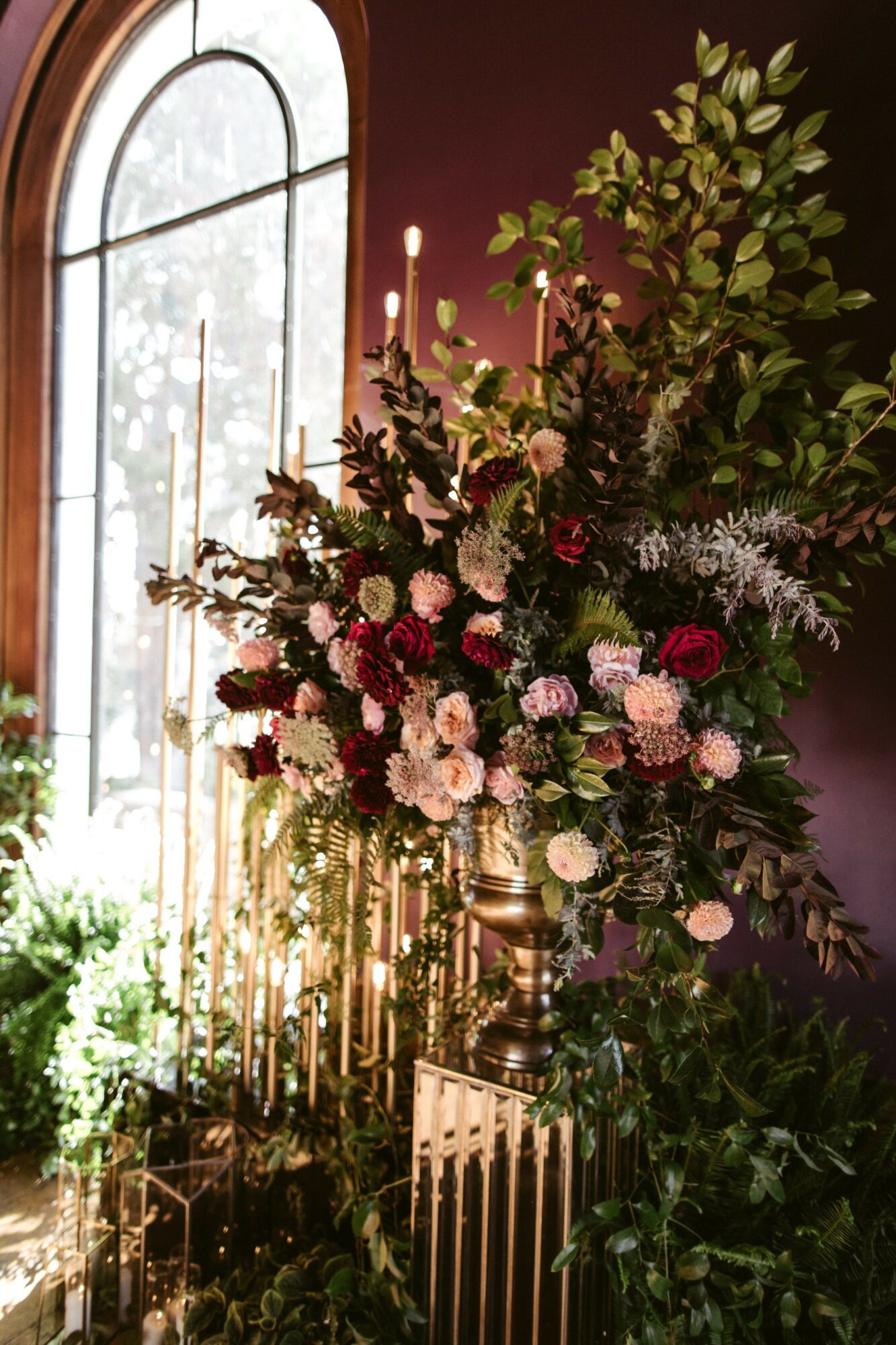 Large floral arrangement with pink, white, and purple flowers, green foliage, in a gold vase, near a window with sunlight.