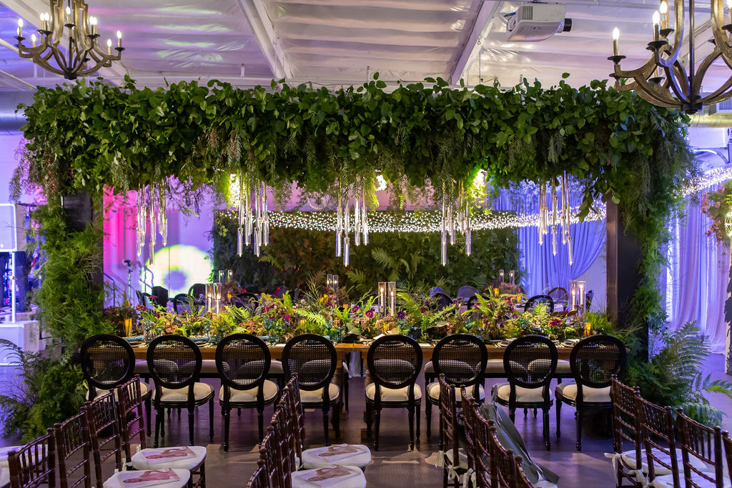Elegant banquet table decorated with greenery, flowers, and hanging crystal ornaments, set in a decorated event space with purple lighting.